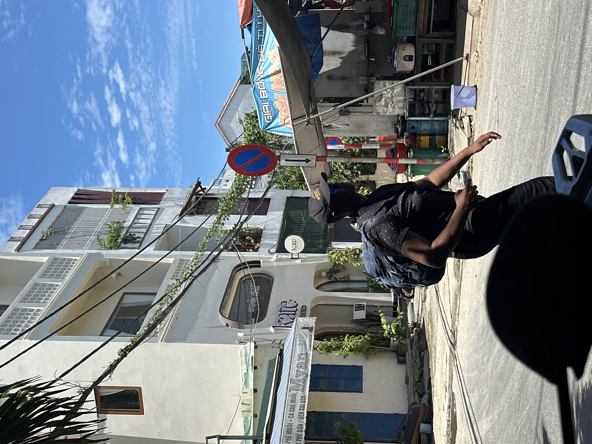 The image shows a street scene in Đà Nẵng, Vietnam, on a sunny day with a blue sky and scattered clouds. A person wearing a black t-shirt, black pants, a black baseball cap, and sunglasses, carrying a blue backpack, is walking away from the camera on the right side of the frame. They are walking on a paved street.
In the background, a multi-story building with white and beige walls is visible. The building has several windows and balconies with greenery. A sign above an arched entrance reads "kate COFFEE & TEA". To the left of this building, another smaller building has a blue door and a striped awning with the text "Myan". Overhead, a tangle of electrical wires crisscrosses the scene. Further down the street, there is a street stall with a blue tarp and a banner that reads "GIẢI BÓNG ĐÁ TỰ DO". Traffic signs, including a red and blue circular sign with a diagonal line, are also present. Various containers and equipment are visible near the street stall. The foreground is dominated by the dark shadow of a motorcycle.