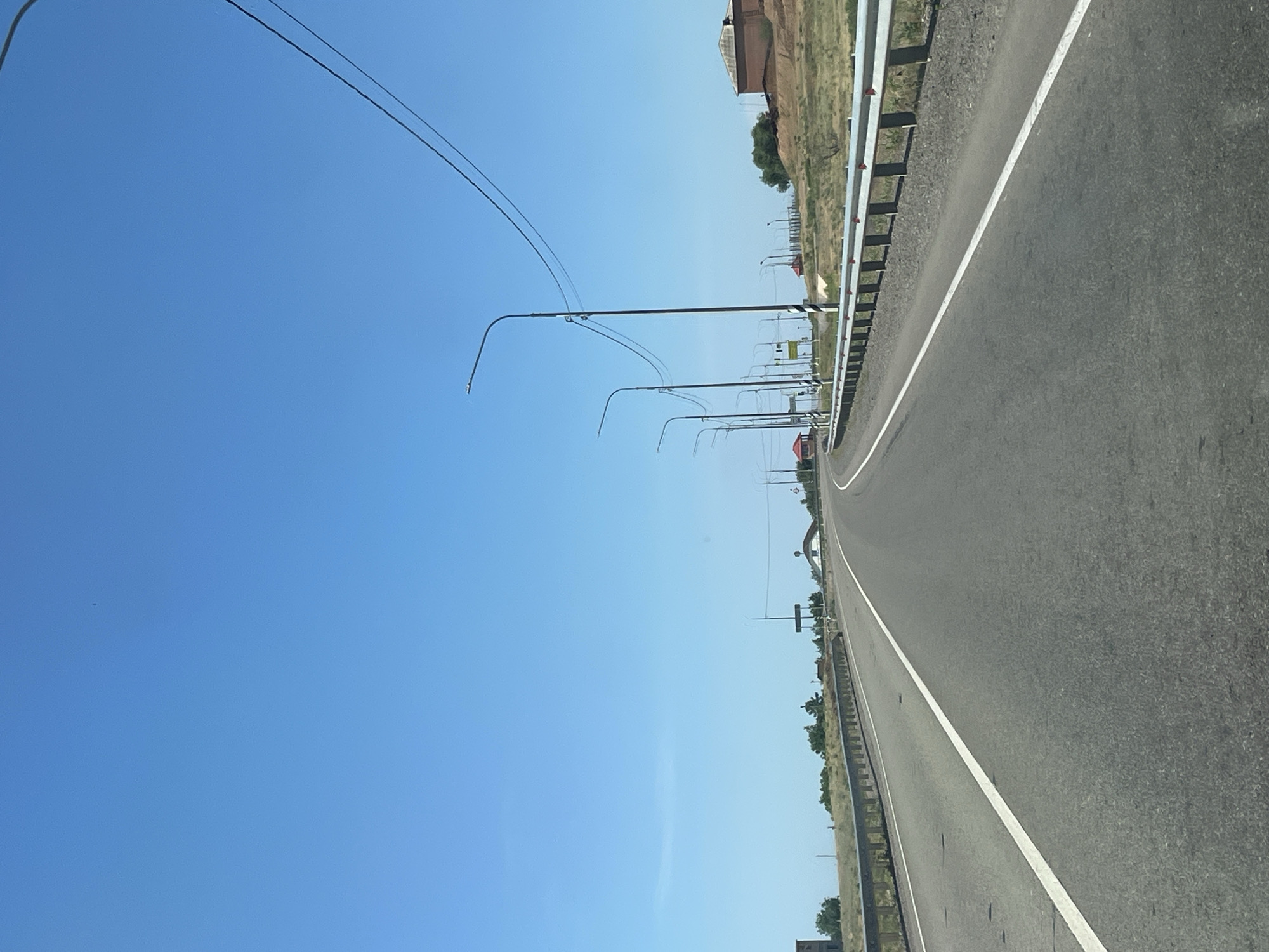 The image shows a two-lane asphalt road with a white line dividing the lanes, seen from the perspective of a vehicle driving on it. The road curves slightly to the right. A metal guardrail with red reflectors runs along the right side of the road. Beyond the guardrail, the landscape is dry and hilly, with sparse vegetation and some buildings visible in the distance. There are several streetlights lining the road, and power lines are strung between poles. The sky is clear and blue. The scene is consistent with a road on the outskirts of Bakhtemir, Russia, likely on a sunny day. No people or animals are visible in the image.