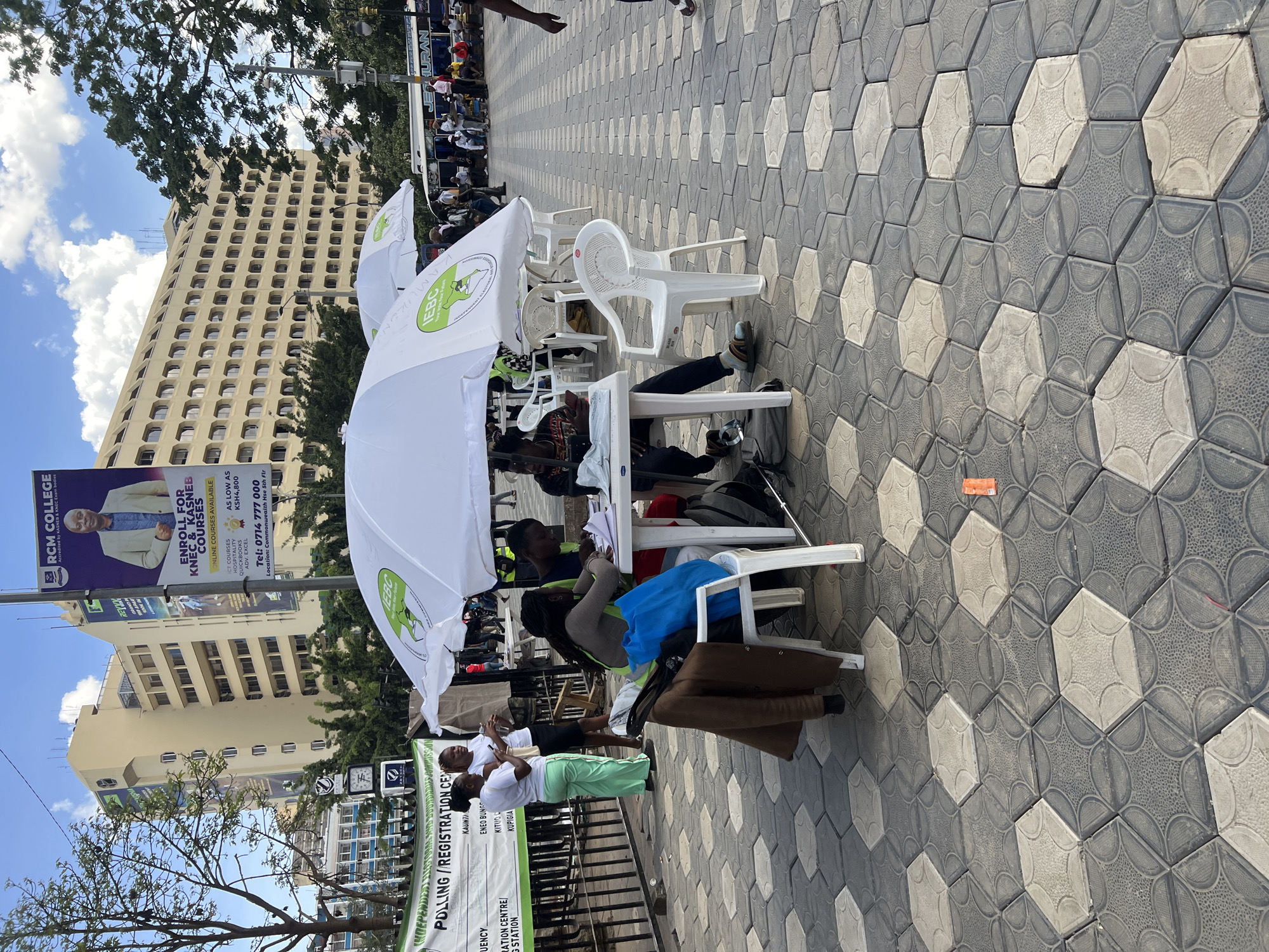 The image depicts an outdoor scene in Nairobi, Kenya, likely a voter registration or polling center. Several people are sitting at white plastic tables under white umbrellas bearing the IEBC (Independent Electoral and Boundaries Commission) logo. These are registration clerks assisting people with voter registration. Two registration officials in matching white shirts and light green trousers are standing. A large billboard for RCM College, advertising KASNEB & KNEC courses, stands in the background. The pavement is patterned with hexagonal tiles. In the background is a tall building. Buses are also visible in the background.