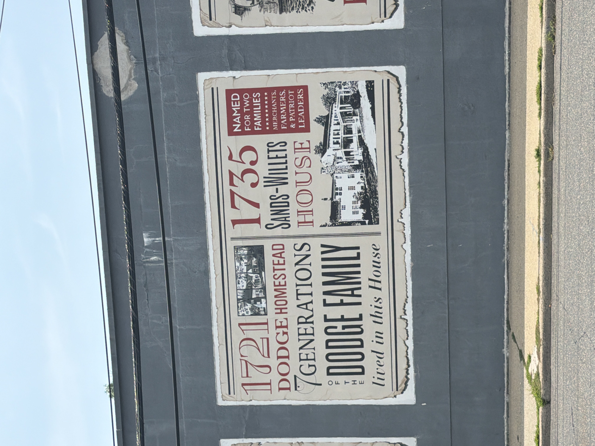 The image shows a mural on the exterior of a building in Port Washington, New York. The mural is divided into sections, with the central and largest section featuring text and an illustration related to the Dodge Homestead. The text reads: "1721 DODGE HOMESTEAD 7 GENERATIONS OF THE DODGE FAMILY lived in this House". To the right of this section, another section of the mural is visible with text that reads: "1735 SANDS-WILLETS HOUSE NAMED FOR TWO FAMILIES ********* MERCHANTS, FARMERS, & PATRIOT LEADERS". The illustration in this section depicts a large house. The mural is painted on a dark gray wall. A sidewalk and street are visible in the foreground, with some grass growing along the edge of the sidewalk. The sky is visible in the background, with some clouds. The mural appears to be old and somewhat weathered, with torn edges visible on the painted sections.
