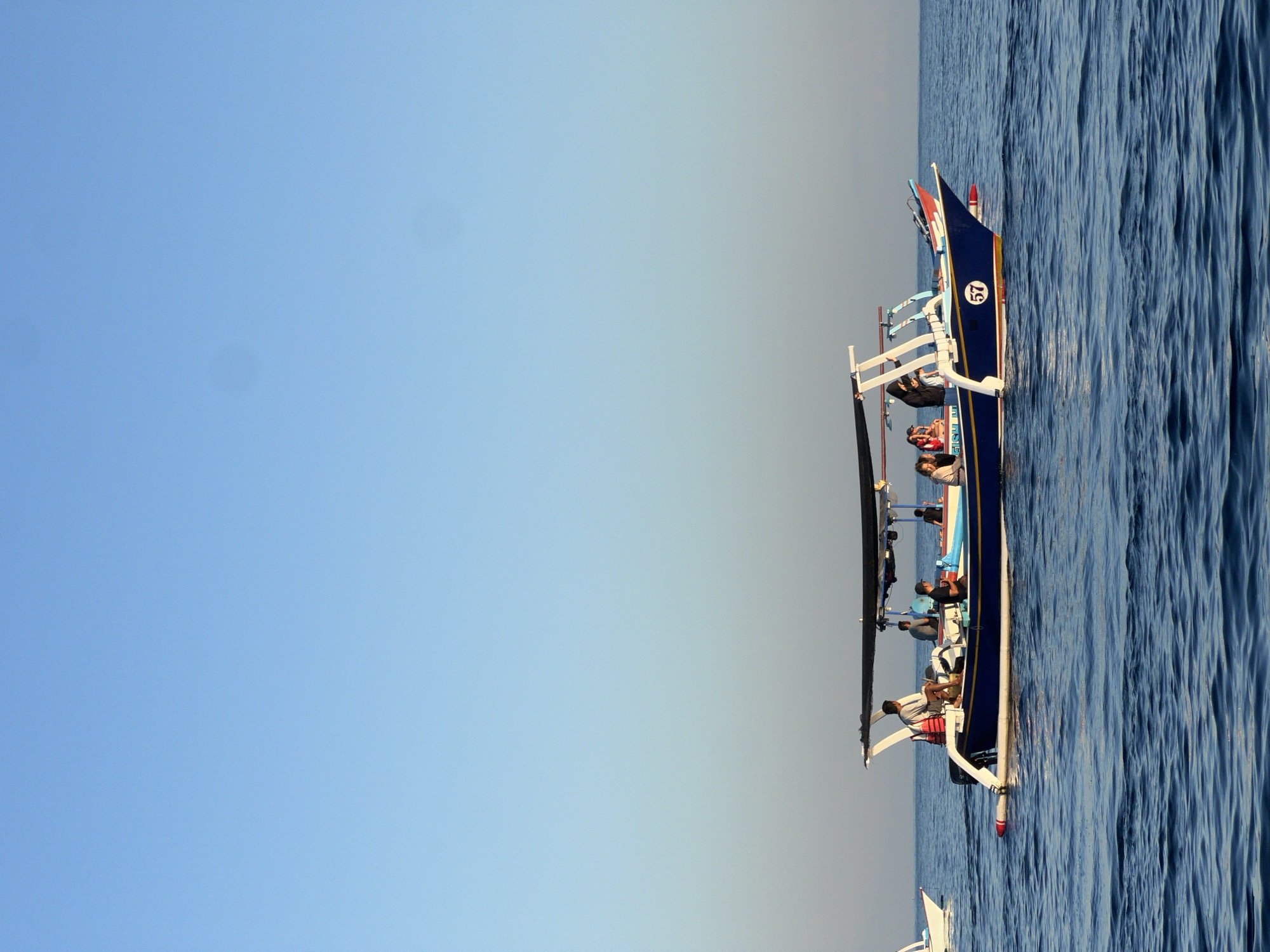 An elevated, eye-level shot captures a deep blue ocean under a clear, pale blue sky. In the middle ground, a narrow, blue and yellow boat with a black awning navigates the choppy water. The boat, numbered "57" on its side, carries several individuals, including a man in a white shirt sitting at the stern and others seated or standing under the awning. Another smaller white boat is partially visible to the left. The scene is peaceful and suggests a calm day at sea. The location is unknown.