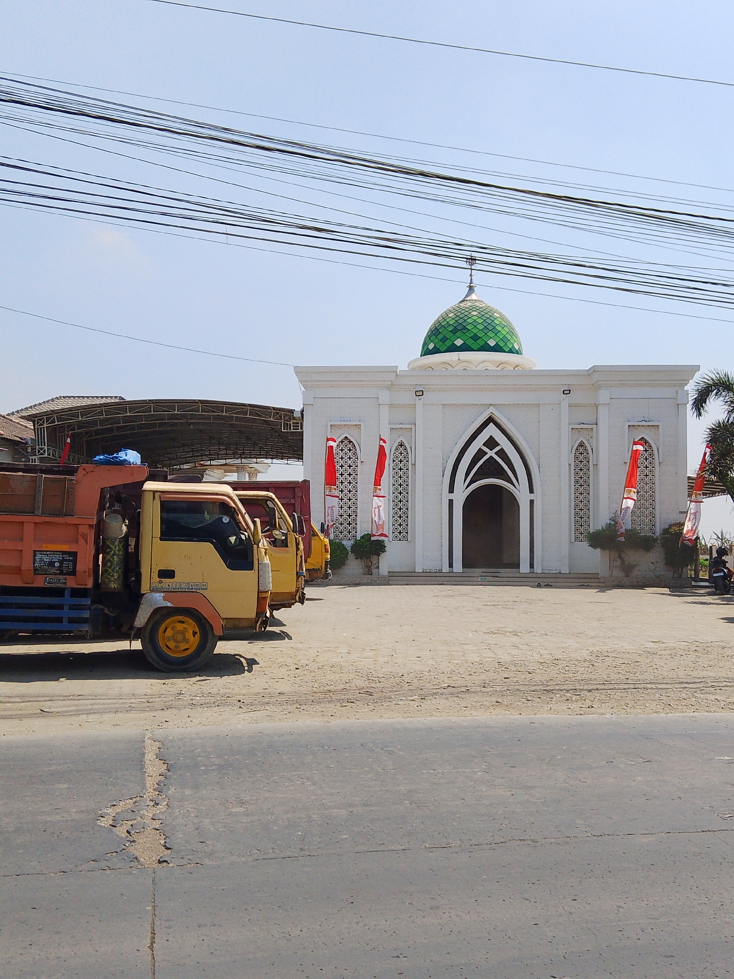 The image depicts a bright, sunny day with a clear blue sky. In the foreground, a paved road with some cracks is visible. Behind the road, a sandy lot is present, with several trucks parked in it. The trucks appear to be utility vehicles, possibly for transport or construction, with the closest one being a yellow and orange flatbed truck.
Further back, a white mosque with a green domed roof stands prominently. The mosque features an arched entrance and decorative latticework on its windows, flanked by tall, red and white Indonesian flags. There are also some bushes and a palm tree near the mosque. A covered structure with a metal roof is located to the left of the mosque, behind the trucks. The scene suggests an outdoor setting in a moderately developed area, potentially a town or city outskirts, given the presence of both the mosque and the commercial vehicles. The overall impression is one of a functional, everyday scene under good weather conditions. The location is identified as Bengkal, Indonesia.