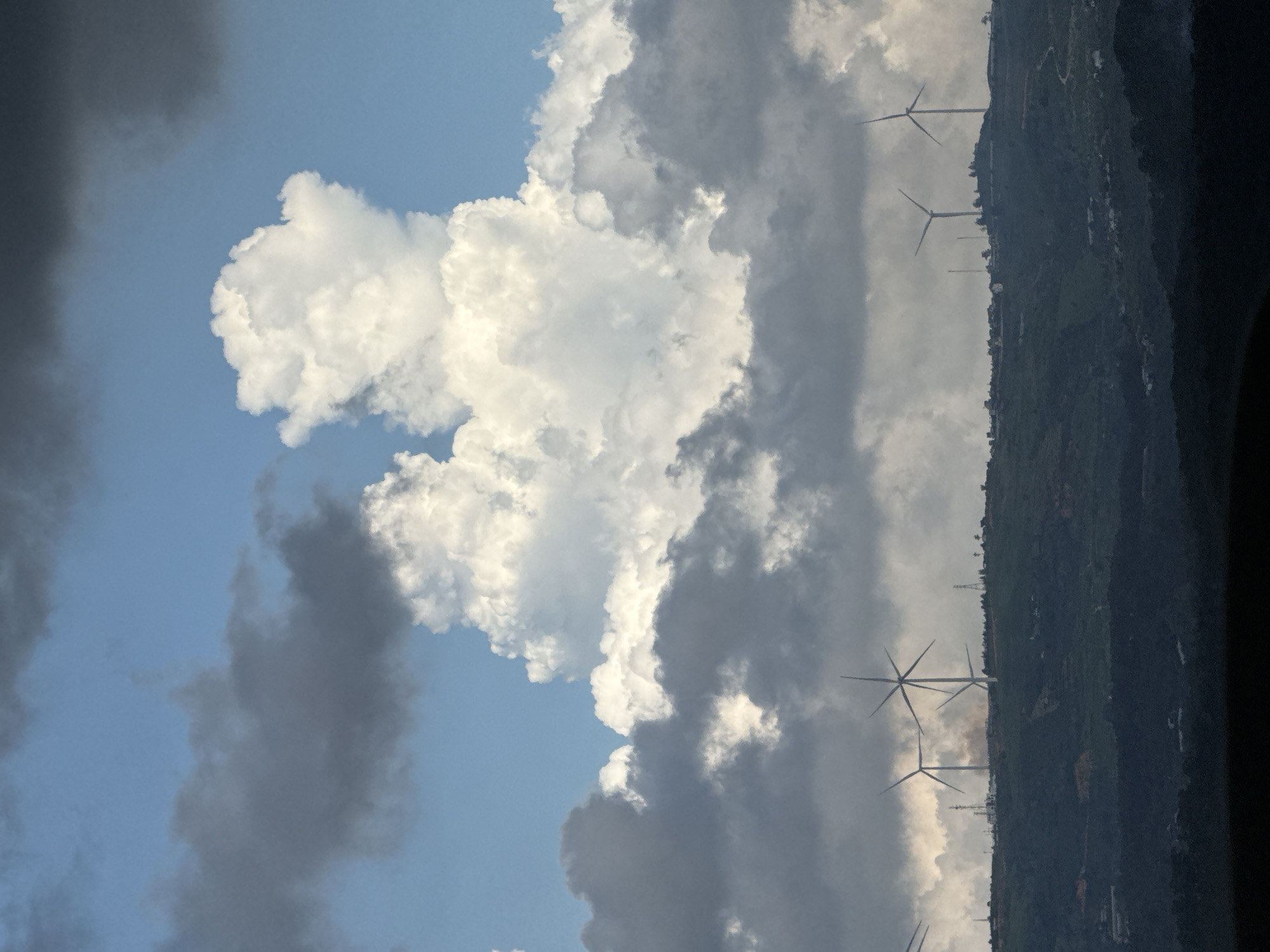 The image depicts a scenic view of rolling hills with several wind turbines positioned on the crest of the landscape. The sky is dramatic, filled with a mix of dark, heavy clouds and bright, puffy cumulus clouds, suggesting changing weather conditions. In the foreground, the rolling hills are covered in dense, dark green vegetation, with some hints of structures and possibly roads visible on their slopes. The wind turbines, with their tall white towers and rotating blades, are silhouetted against the sky and the clouds. The overall scene captures a sense of vastness and the power of nature, with the wind turbines representing a human intervention in this natural environment. The specific location is identified as Đa Lộc, Vietnam.