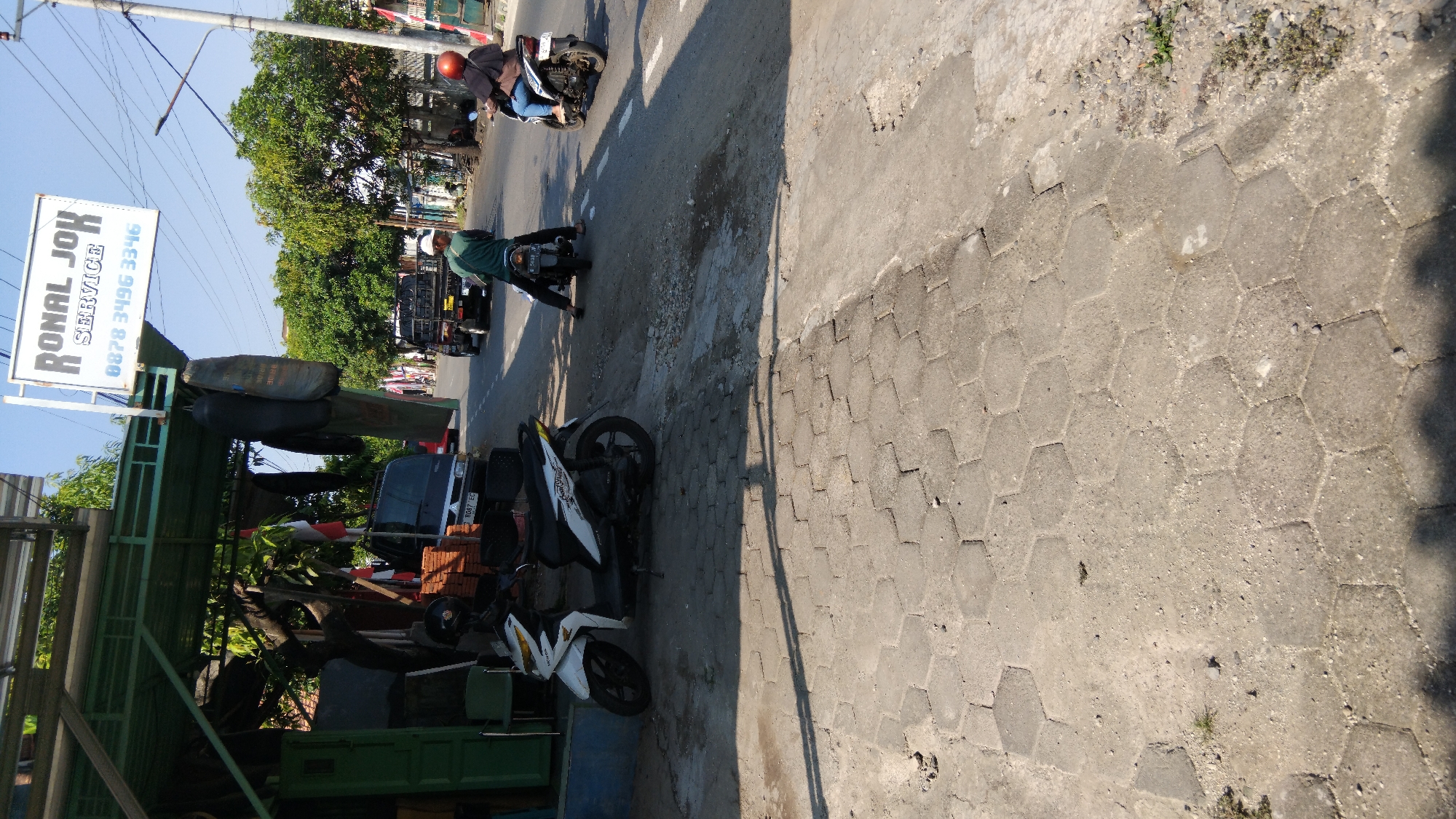In the city of Proyonanggan Tengah, Indonesia, a street scene unfolds under a bright, clear sky. The foreground is dominated by a textured, hexagonal-patterned concrete walkway. To the left, a white Honda scooter is parked, its kickstand down. Behind the scooter, a covered open-air structure with a green metal roof is visible, housing various items including what appear to be spare tires hanging from the roof and a red and white Indonesian flag. A dark-colored van is parked under the shelter. Further down the street, two individuals are riding motorcycles. One person, wearing a green shirt and a white helmet, is riding a motorcycle in the middle of the road, facing away from the camera. Another person, wearing a dark shirt and an orange helmet, is riding a motorcycle on the right side of the road, also facing away. Several vehicles, including a black pickup truck and another dark-colored pickup truck, are also present on the road. Trees line the sides of the street, and a utility pole with a street light stands on the right. A white sign with the words "RONAL JOK SERVICE" and a phone number is mounted above the covered structure.