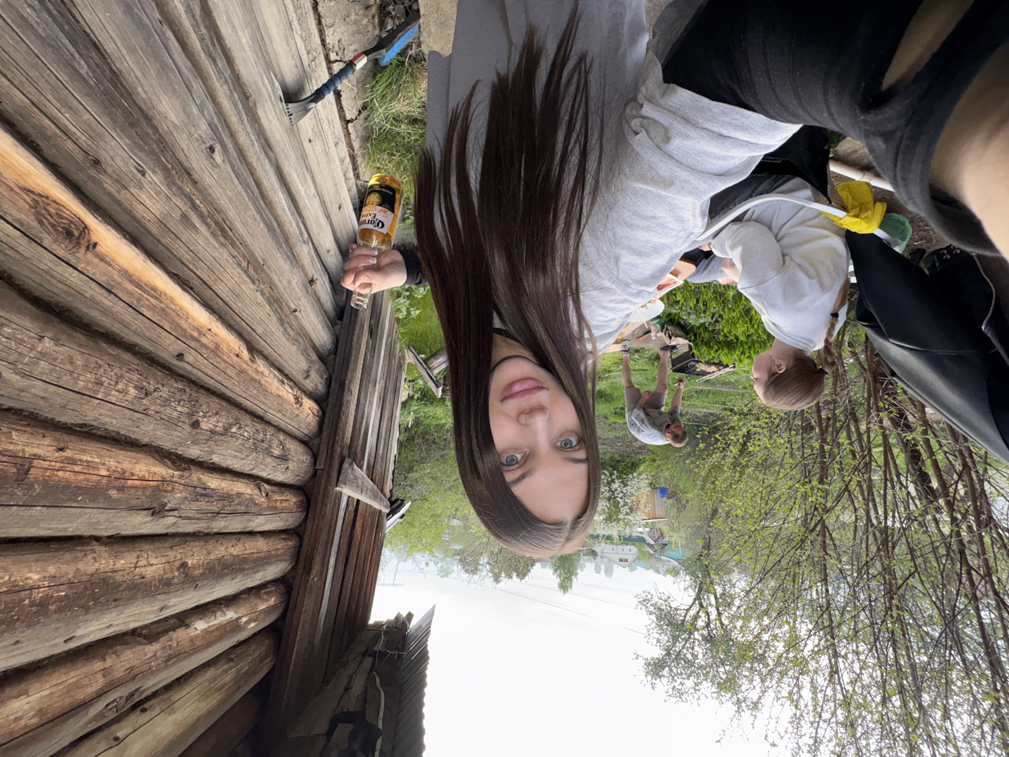 The image shows an outdoor scene in the city of Chaikovsky, Russia, with a young woman as the central subject. She is holding a Corona beer bottle in her right hand. She appears to be at a backyard gathering near a wooden log cabin, with other individuals visible in the background tending to a grill. The surrounding environment includes trees and other vegetation characteristic of early spring. In the far background, a few houses of the city are visible.