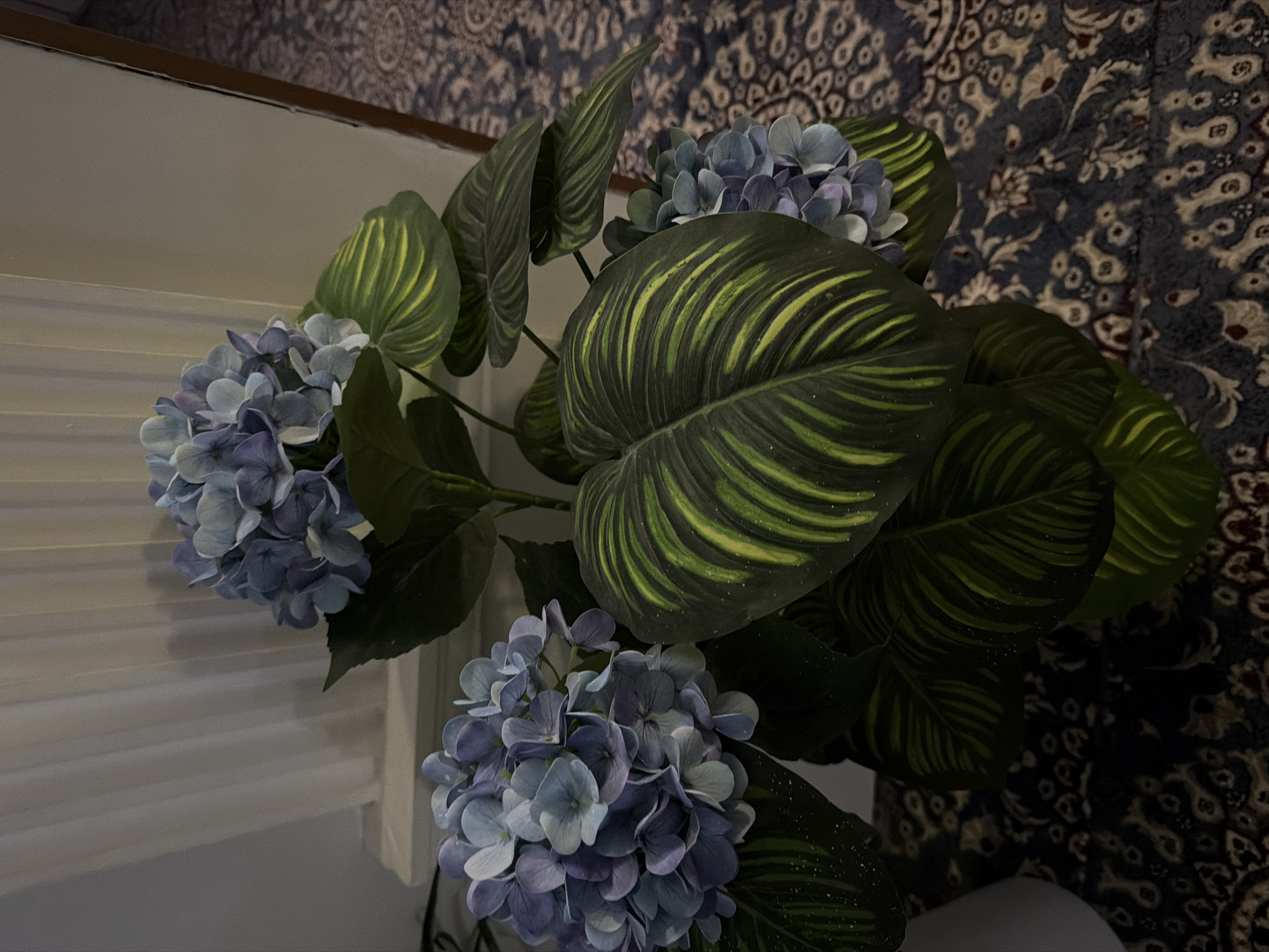 The image shows a potted artificial plant with blue hydrangeas and large, dark green leaves with yellow stripes. The plant is placed on a dark, patterned carpet. In the background, there is a white pleated curtain or screen and a light-colored wall. The scene appears to be indoors, likely in a residential setting in the city of Khumsan, Uzbekistan. There are no people or animals visible in the image. The lighting is dim, suggesting it might be evening or an overcast day.