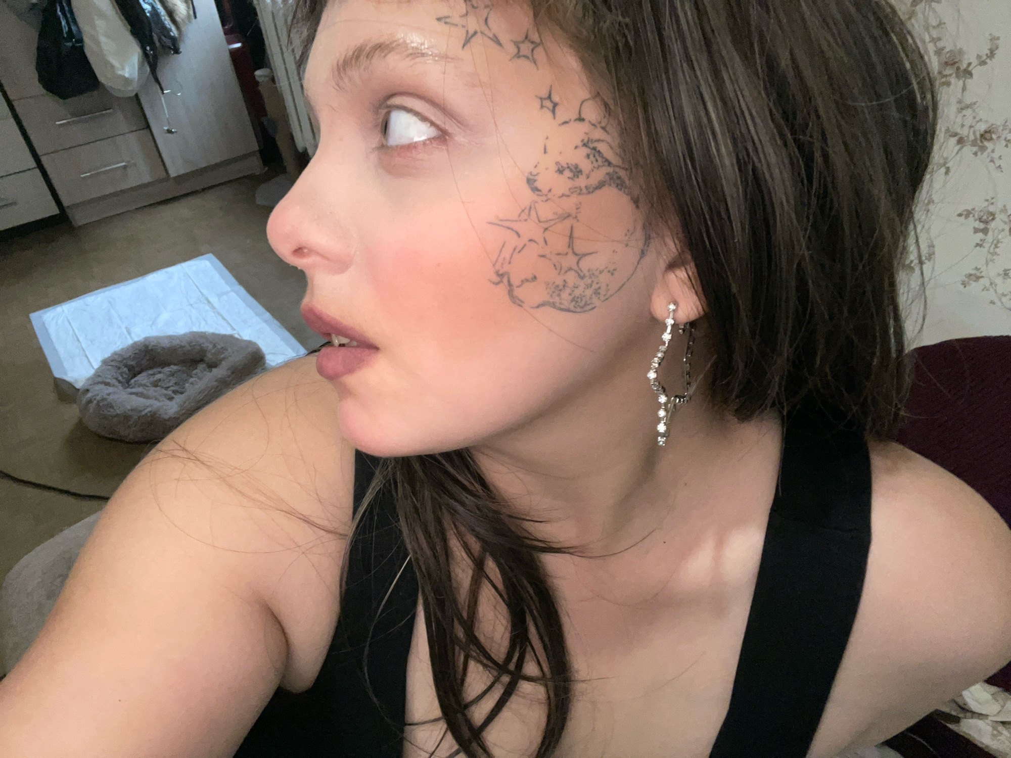 The image shows a close-up, side profile of a young woman's face, with a tattoo of a bear surrounded by stars on her cheek. She is looking to her left, with her mouth slightly open. She is wearing a black tank top and a sparkly dangle earring. The background is blurred, but appears to be an indoor setting with furniture and a floral wallpaper. There is no indication of the city of Lipetsk, Russia in the image.