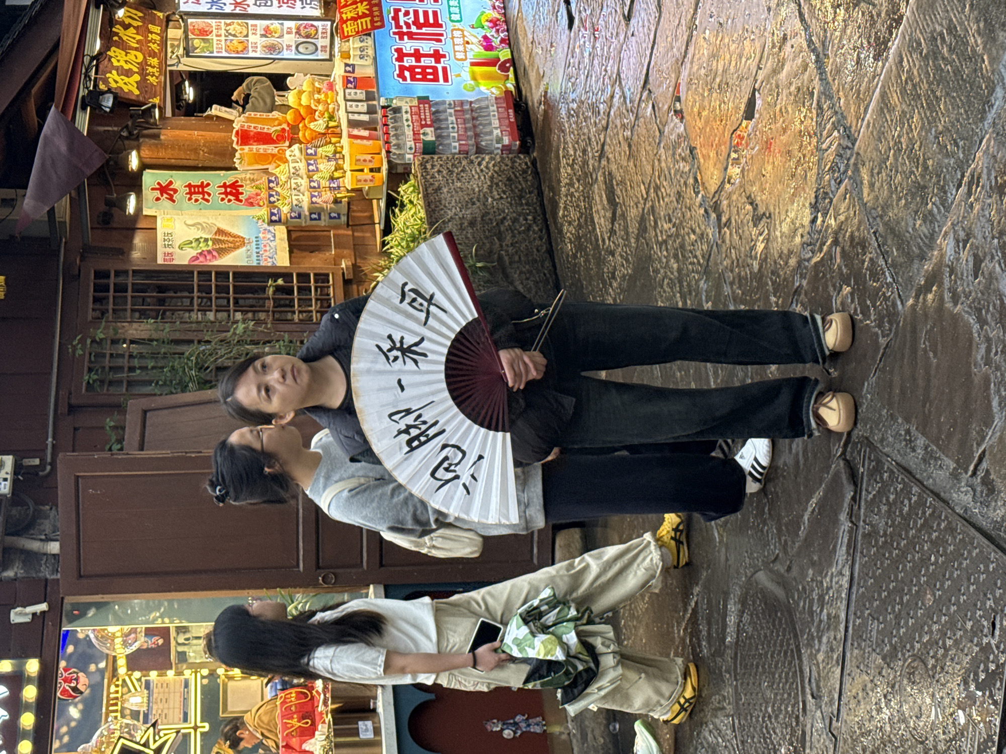 This image captures a vibrant street scene in Shapingba District, China. The scene is a street lined with food vendors. Three women stand in the foreground: one with long black hair holds a phone and a patterned bag; another woman next to her wears glasses and a light-colored sweater; a third woman holds a Chinese fan. The environment is a bustling street with various food stalls, their signs displaying information in Chinese. The ground appears to be wet.