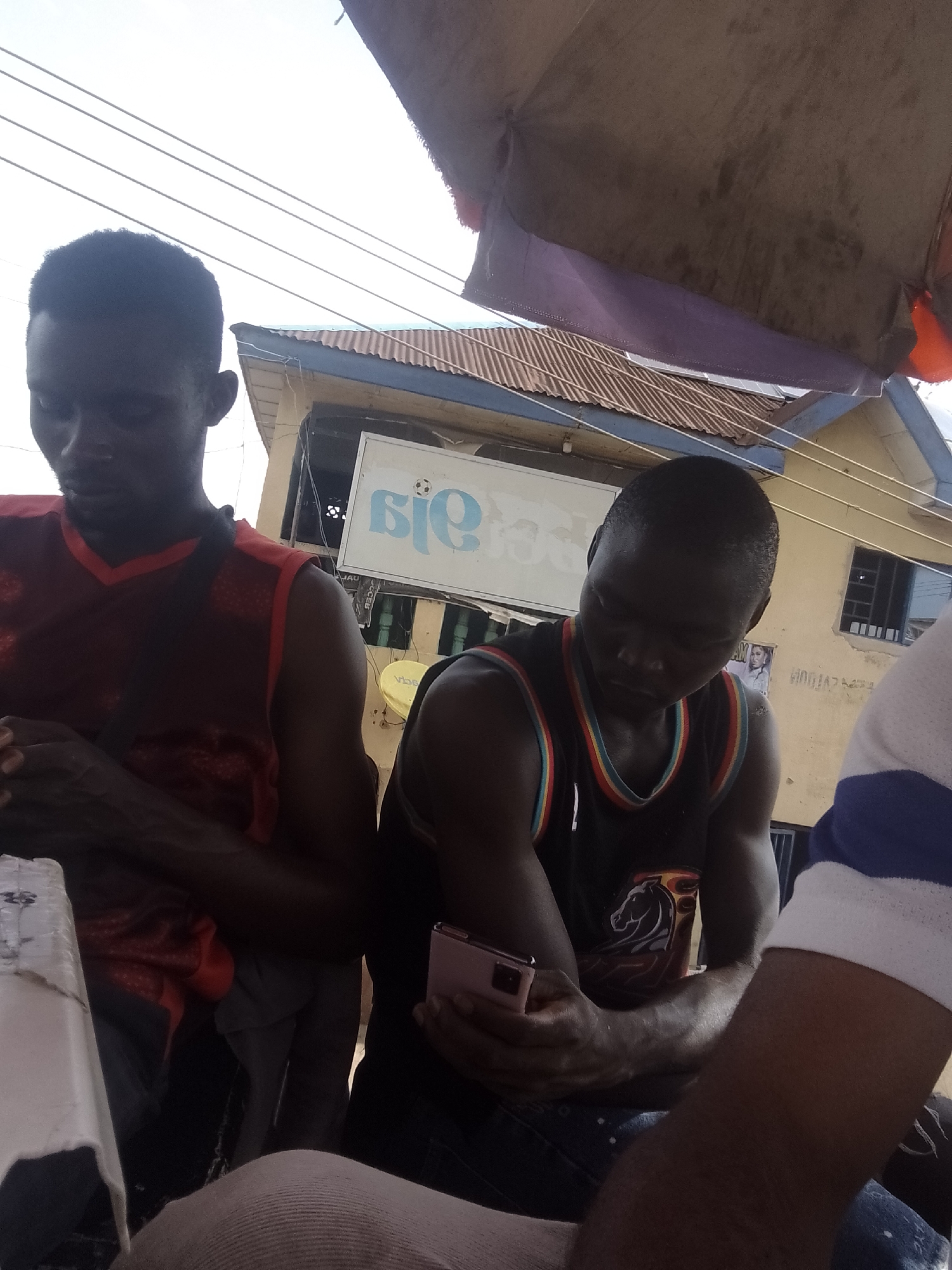 The image shows a close-up view of two young Black men sitting at a table outdoors. Both men are wearing sleeveless athletic shirts. The man on the left has on a red shirt with a dark pattern, and the man on the right is wearing a black shirt with colorful trim and a graphic of a horse. The man on the right is holding a pink smartphone and looking down at it. A third person's arm, wearing a blue and white striped sleeve, is visible on the far right of the frame. In the background, there is a building with a corrugated metal roof and a yellow facade. A sign with blue lettering and a soccer ball is mounted on the building. Overhead, wires and parts of an umbrella or awning are visible. The scene appears to be in a casual outdoor setting, possibly a street-side stall or eatery.