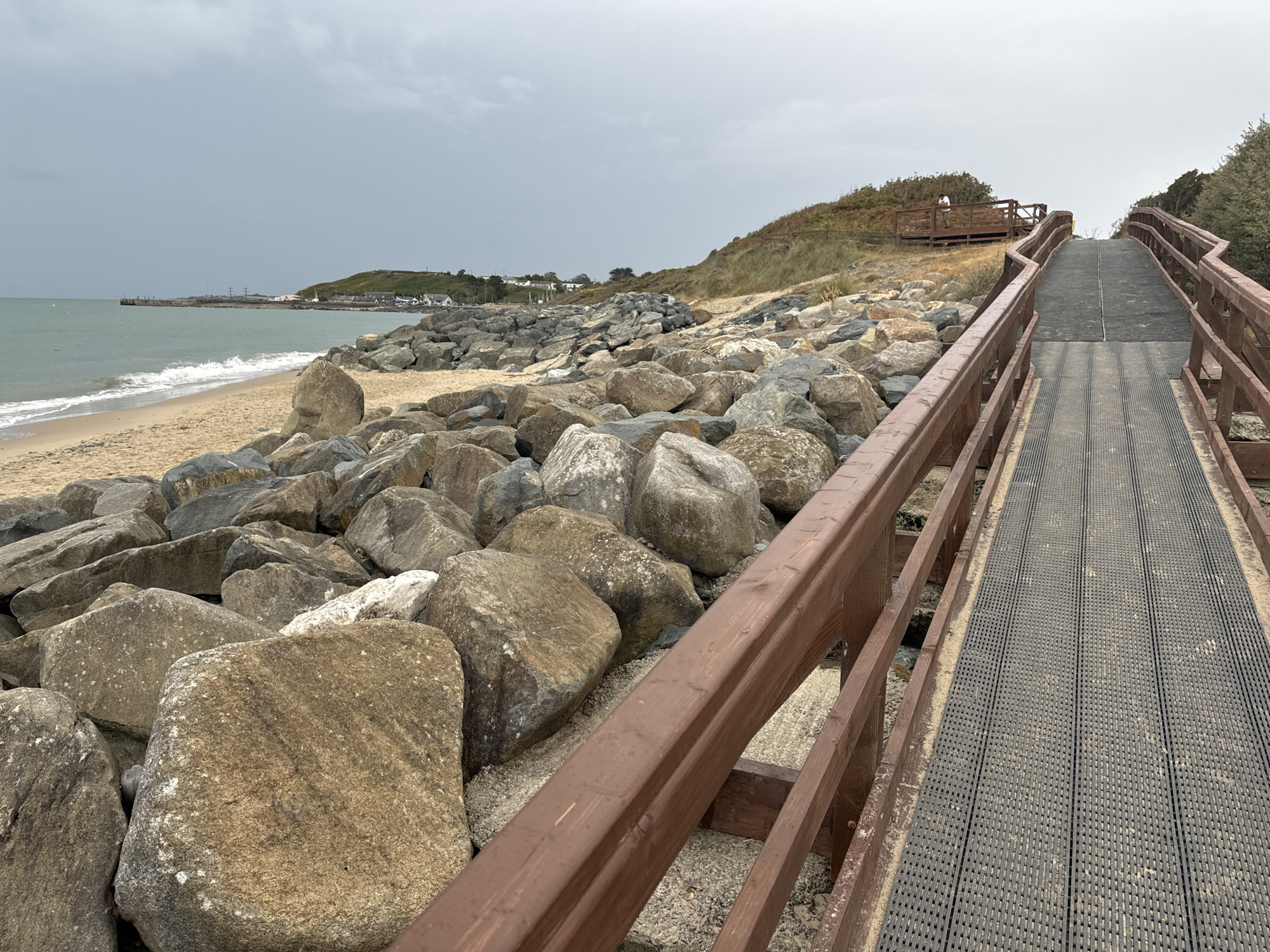 The image shows a wooden boardwalk with dark grey grated decking, curving upwards along a rocky shoreline. The boardwalk has brown wooden railings. To the left of the boardwalk is a beach composed of sand and large, rough rocks. Beyond the rocks, the sea is visible with waves breaking on the shore. In the distance, across the water, there is a pier with some structures and buildings on a grassy hill. The sky is overcast and grey. The scene is located in Courtown, Ireland, along the coast.