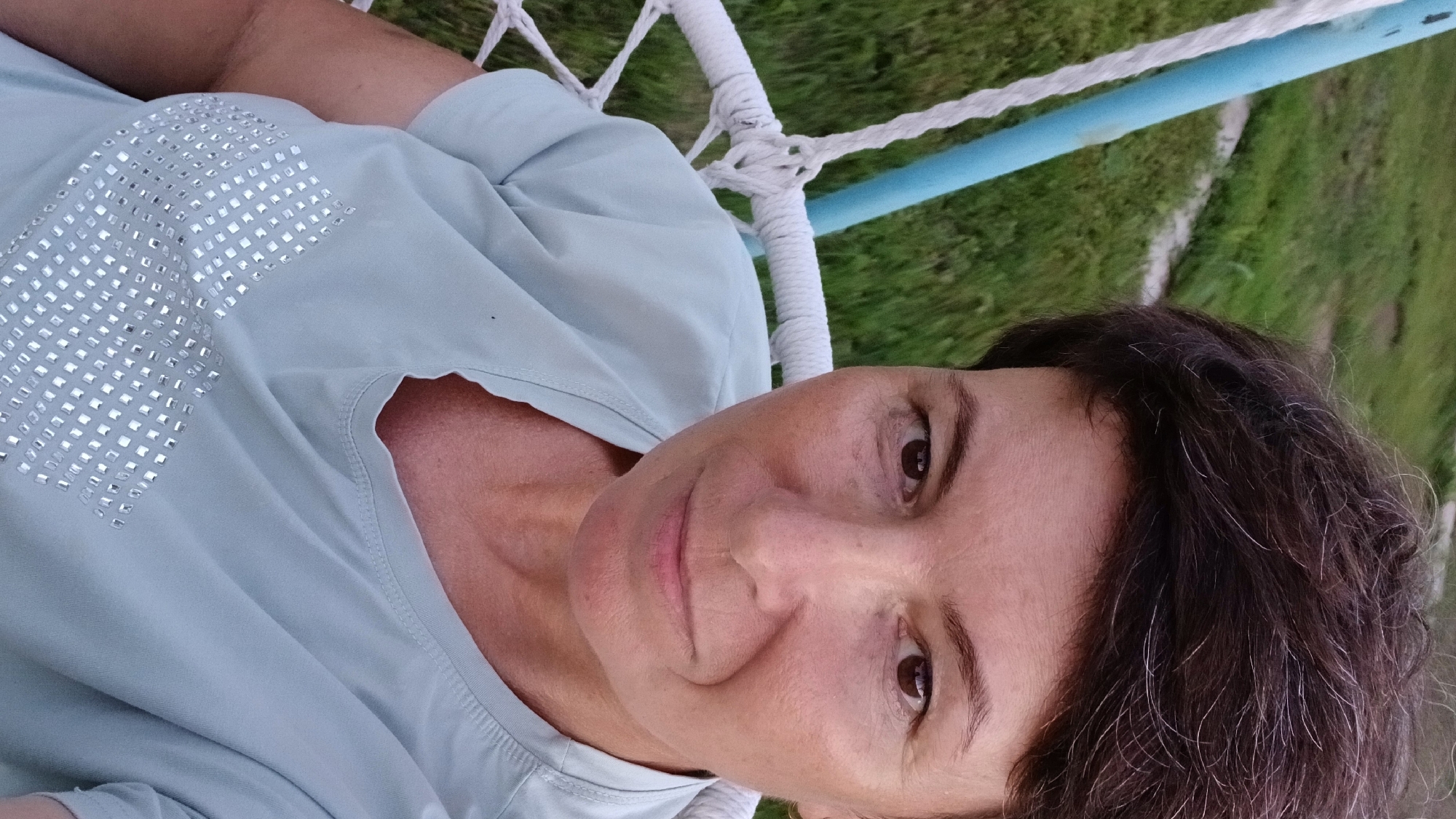 In Nadezhdenka, Russia, a woman with short, dark hair is captured in a selfie. She is seated in a white hammock chair with a blue support pole visible to her left. The background consists of green grass and possibly some concrete structures. The woman is wearing a light blue t-shirt adorned with a pattern of small, reflective squares across the chest. Her gaze is directed towards the camera. The lighting suggests it is daytime, possibly late afternoon or early evening.