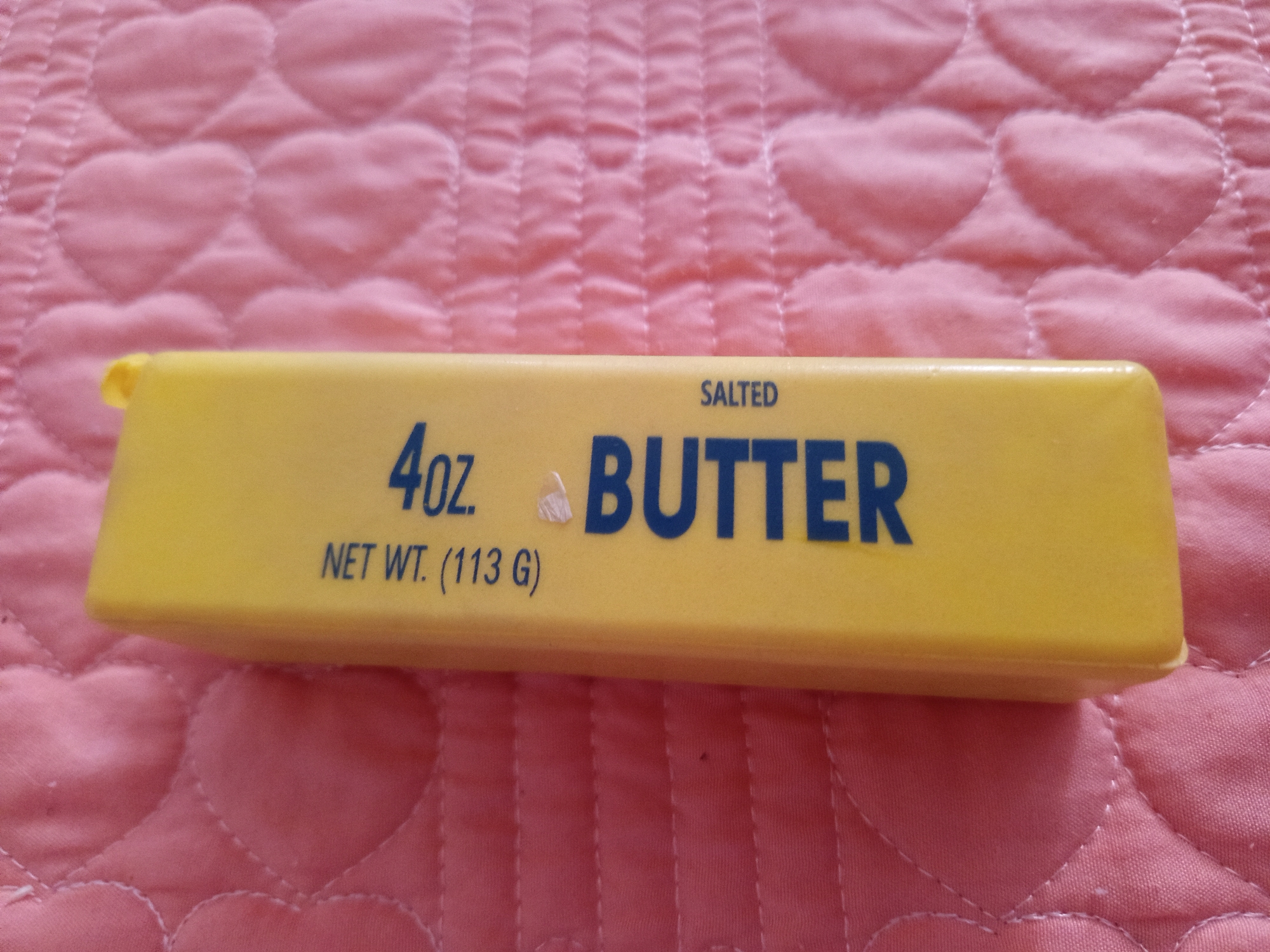 A stick of yellow butter with the words "SALTED BUTTER" and "4oz. NET WT. (113 G)" printed on it lies horizontally on a pink quilted surface. The surface is textured with stitched heart shapes. There is no information available about the city of Talapty, Kazakhstan.