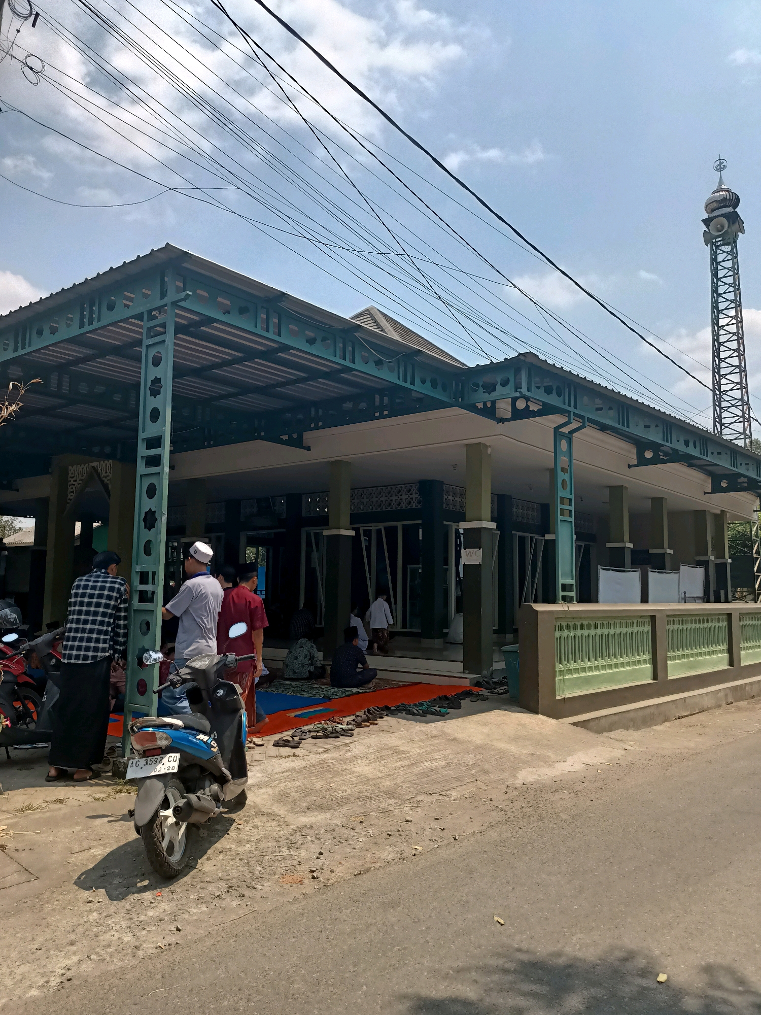 The image captures a street-level view of a mosque in the city of Badas, Indonesia. The mosque features a prominent green metal roof structure supported by pillars, with a wider, more open area in the foreground. A tall, lattice-work tower, likely a minaret, stands to the right, topped with a dome and decorative elements, and equipped with loudspeakers. Several individuals, some wearing traditional Islamic attire like sarongs and peci (caps), are visible around the mosque. Some are standing near motorcycles parked in the foreground, while others are seated on prayer mats inside the mosque's covered veranda. The ground is a mix of concrete and asphalt, with a green decorative fence bordering the mosque's property. Overhead, a tangle of power lines crisscrosses the sky, suggesting an urban or semi-urban environment. The atmosphere appears calm, possibly indicating a prayer gathering.