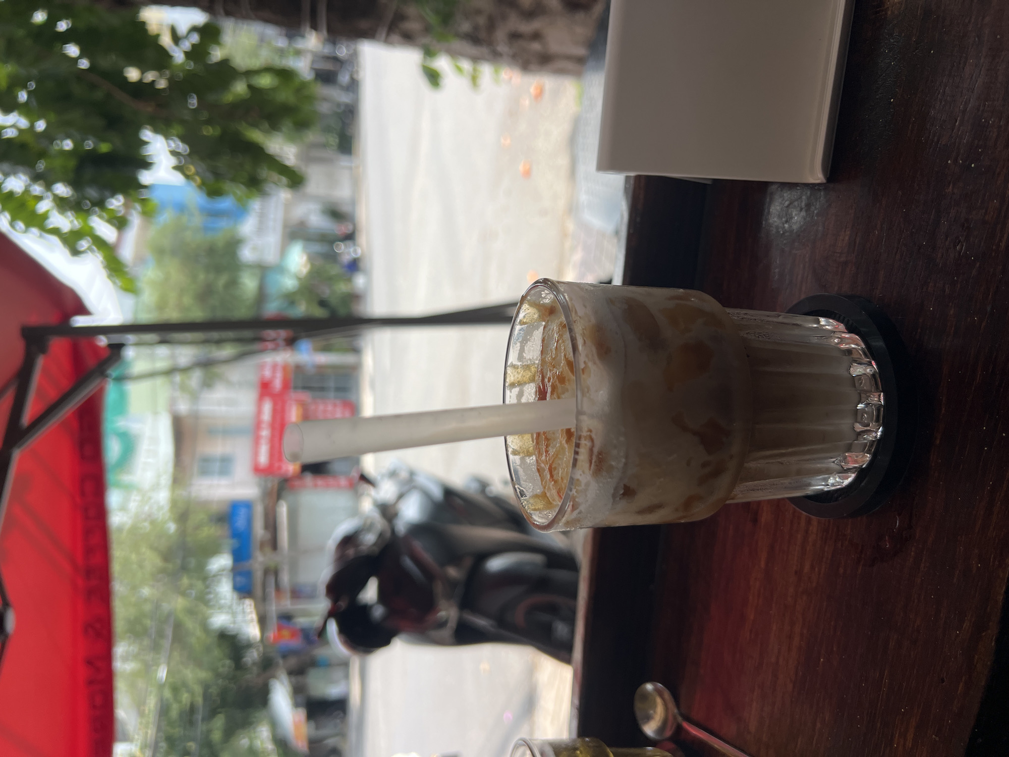 The image captures a glass of iced coffee or tea with a white straw, positioned on a dark, polished wooden table, likely within a cafe. The beverage contains ice and appears to be a milky or creamy mix with a brownish liquid, possibly caramel or coffee, swirled within. The glass rests on a black coaster.
Through a large window or opening, a bustling outdoor street scene is visible, mostly out of focus. Green trees and foliage are prominent, indicating an urban setting with natural elements. Beyond the trees, buildings line the street, with some signs and architectural details discernable. Several motorcycles, typical of Vietnamese streetscapes, are parked or moving on the road. A bright red awning, likely belonging to the establishment, is visible on the left, displaying mirrored white text that reads "CÀ PHÊ & VIỆT", confirming the setting as a Vietnamese cafe.
The scene is illuminated by natural daylight, suggesting it is daytime. The view from inside the cafe offers a glimpse into the dynamic street life of Nam Nha Trang Ward, Vietnam.