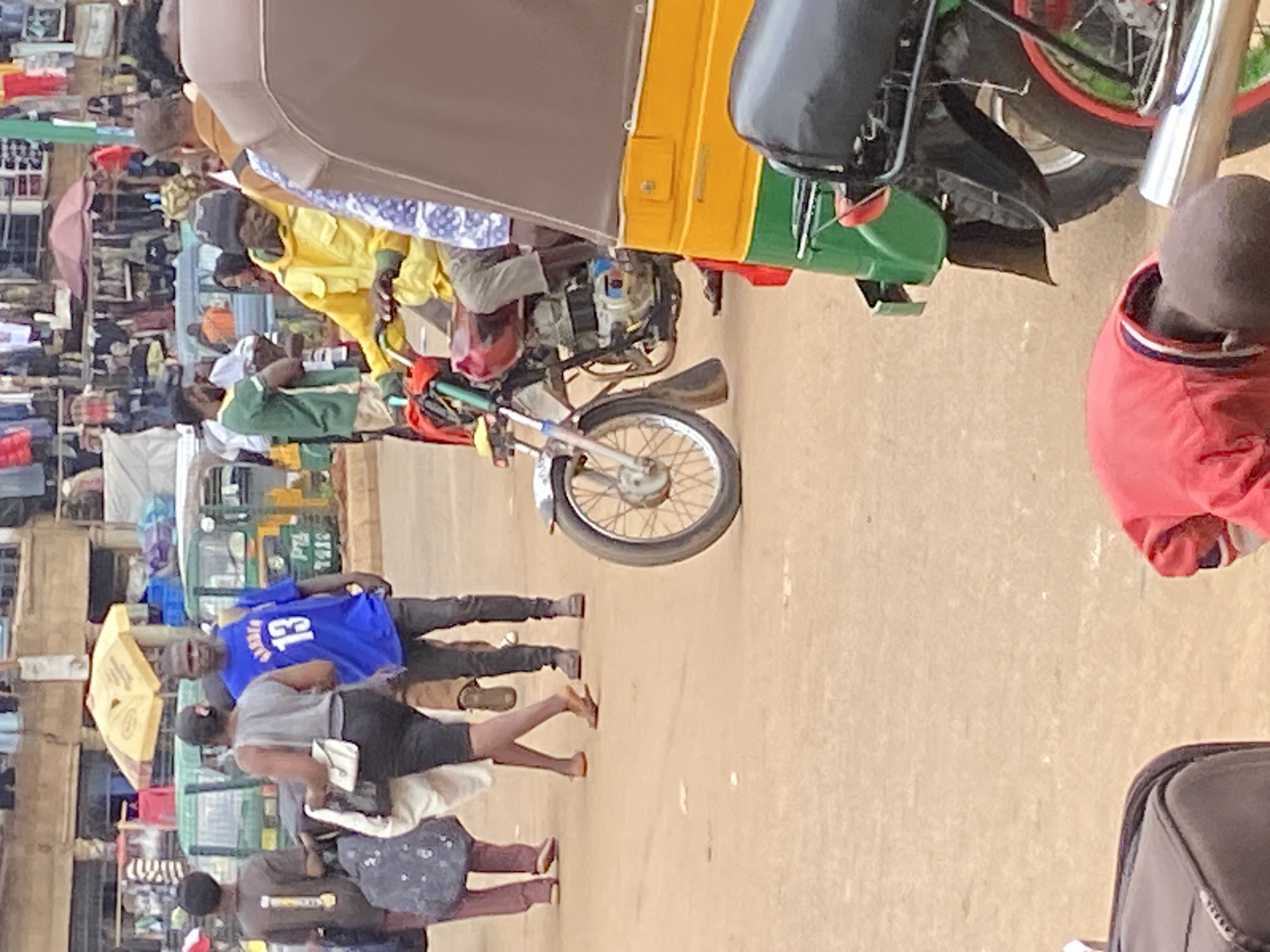 In Bukuru, Nigeria, a busy street scene unfolds. In the foreground, a motorcycle with a prominent engine and wheel, and a yellow, green, and brown auto-rickshaw are parked. Behind them, a man in a bright yellow jacket is seated on the motorcycle. Several pedestrians are visible: a man in a blue shirt with the number "13" on it, a woman in a grey tank top and black shorts, and another person in a brown shirt. The background reveals a marketplace with stalls displaying clothing and yellow umbrellas, with more auto-rickshaws and people scattered throughout. The ground is a sandy, earthy color.