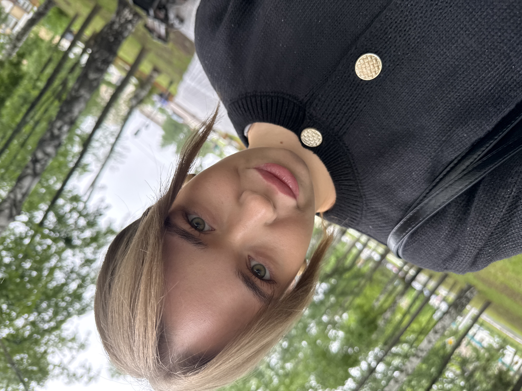 A selfie of a woman outdoors in Chaikovsky, Russia. The woman has light blonde hair, styled in short pigtails, with green eyes and pink lips. She is wearing a black cardigan with gold buttons and a white undershirt. The background shows a blurred outdoor scene with trees and foliage, suggesting a park or green space. The photo appears to have been taken from a low angle, looking up.