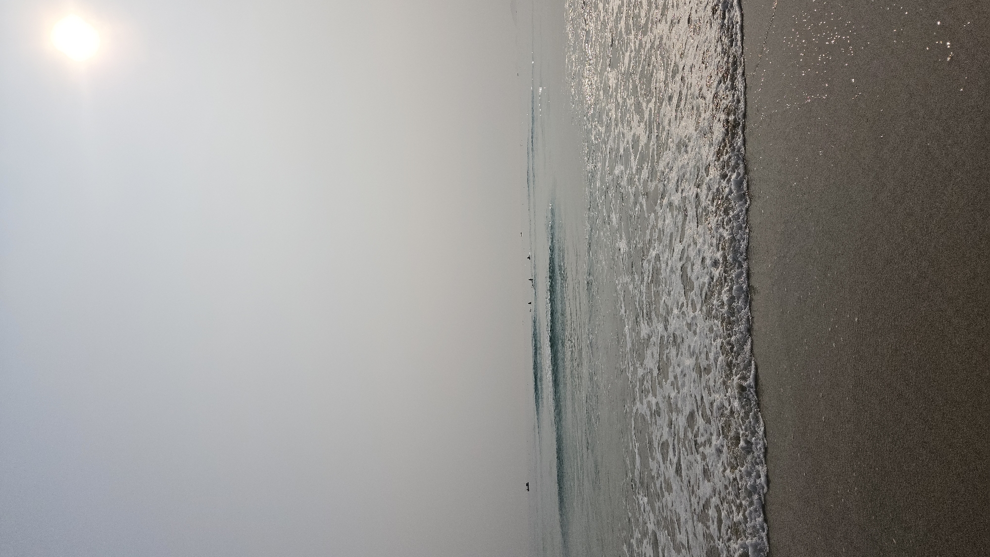 The image shows a beach in Bac Cam Ranh, Vietnam. The sky is overcast with the sun visible as a bright spot in the upper left. The beach spans the right side of the image, transitioning into the ocean, which is a flat, light blue. Along the shoreline, waves are crashing onto the sand. Several indistinct figures appear in the water farther out. The sand is wet and dark.