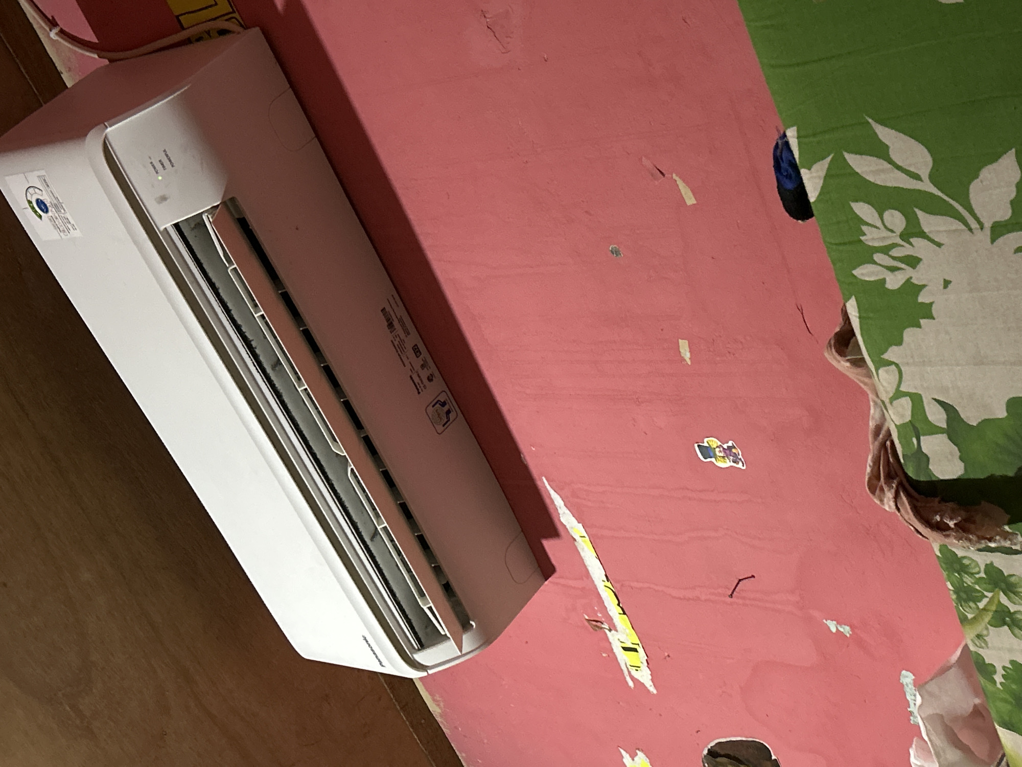 An indoor shot shows a white air conditioning unit mounted on a pink wall. The air conditioner is a Panasonic model. Above the air conditioner is a wooden ceiling. The wall has some peeling paint and yellow and white stickers. In the lower right corner, a green floral patterned cloth is visible. The scene appears to be within a residential setting in Depok, Indonesia.