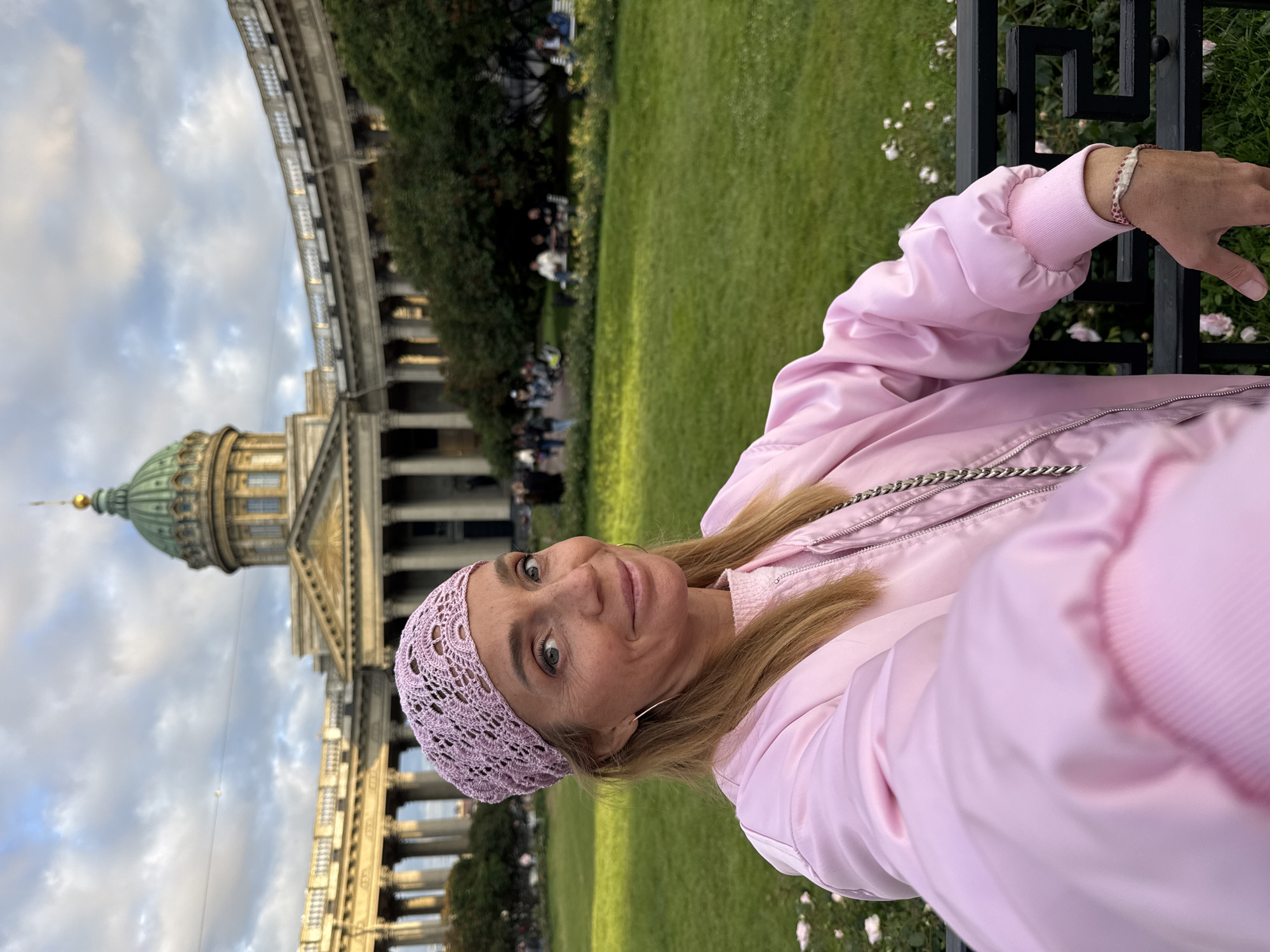 This is a selfie taken by a woman in front of Kazan Cathedral in Saint Petersburg, Russia. She is wearing a pink bomber jacket and a light purple crocheted hat. The dome of the cathedral, a prominent architectural landmark, is visible in the background, along with its colonnade and a lush green lawn. The sky is partly cloudy, suggesting it's daytime. The woman is smiling and looking at the camera, conveying a sense of happiness and enjoyment of her surroundings. The image is oriented vertically, with the woman on the right and the cathedral on the left. There is no visible text in the image.