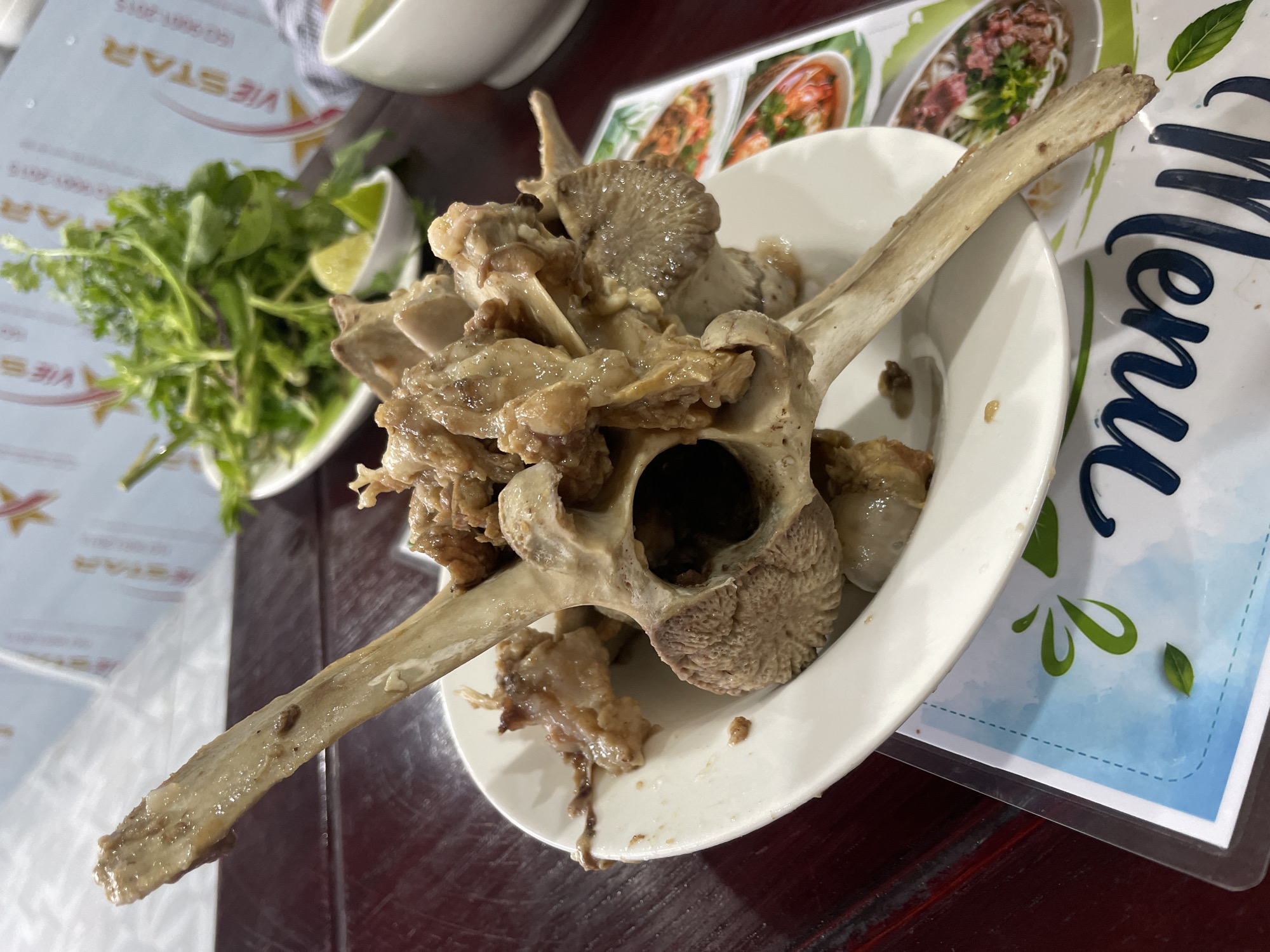 This close-up shot captures a meal being served on a dark wooden table, likely in a restaurant setting. The central subject is a white plate piled with large, cooked animal bones and some tender meat still attached. A prominent, long bone, possibly a rib or a vertebra, spans across the plate, and other smaller bone fragments and meat pieces are visible, suggesting a slow-cooked or stewed dish. One large bone section displays a hollow cavity, possibly from a spine.
To the left, a small white bowl holds a generous portion of fresh green herbs, including what appears to be cilantro, accompanied by a bright green lime wedge, ready to be added to the dish for flavor.
Underneath the plate and side dish, two different menus or placemats are visible. One, upside down, clearly shows the text "VIESTAR" in large, stylized letters, along with some smaller, indistinguishable text. The other placemat, to the right, features a vertically oriented, cursive "Menu" title against a blue and white watercolor-like background with green leaf and splash graphics. Parts of what appear to be food images, possibly pho, are visible on this placemat. The overall scene suggests a casual dining experience in Phu Thuy Ward, Vietnam.