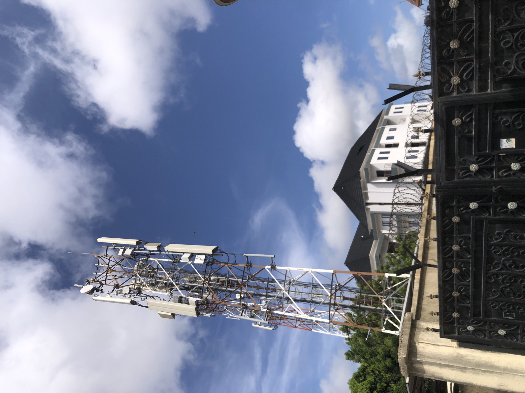 This image, taken in Ado, Nigeria, presents a daytime scene dominated by a tall, lattice communications tower. The tower, constructed from metal, features sections painted white and others with a reddish-brown, possibly rusty, appearance. Numerous antennas, dishes, and cables are affixed to its structure.
In the background, a modern, multi-story white building with a dark, sloped roof is visible. The building has multiple dark-framed windows. Separating the foreground from the building is a robust security fence. This fence is topped with extensive barbed wire. A prominent feature of the fence is a large, dark, ornate metal gate, decorated with recurring circular floral motifs. The base of the fence in the foreground consists of a concrete wall or pillar.
The sky is a vibrant blue, dotted with scattered white cumulus clouds, indicating clear and sunny weather, likely late morning or early afternoon. Green foliage from trees or bushes can be seen near the base of the tower and adjacent to the building. No people are visible in the scene. A small, light-colored sticker or logo is attached to the ornate metal gate, but its text or details are unreadable. The overall setting suggests an urban or suburban environment with a focus on telecommunications infrastructure and property security.