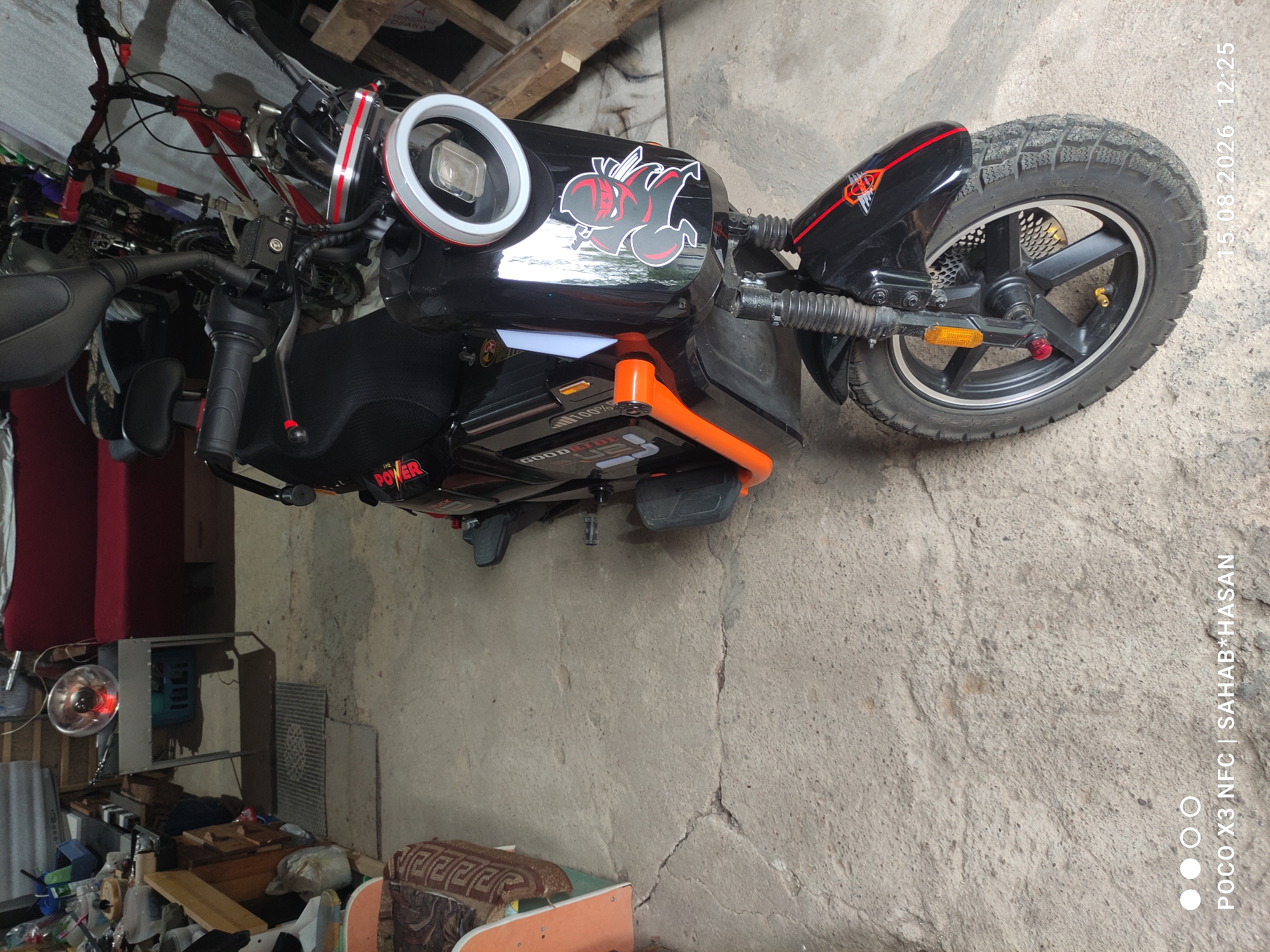 The image shows an electric scooter parked indoors, possibly in a garage or storage area. The scooter is black with orange accents and features a prominent circular headlight with a silver ring. A sticker of a ninja character is visible on the front panel. In the background, other items are stored, including a red sofa, bicycles, and various other objects which are difficult to identify clearly. The floor is made of concrete. The timestamp in the bottom right corner indicates the image was taken on August 15, 2026, at 12:25. The location is identified as Kostroma, Russia.