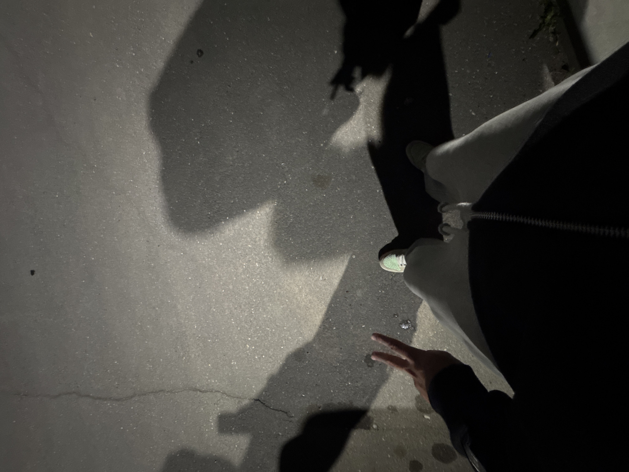 A dimly lit scene, possibly Yerevan, Armenia, judging by contextual cues, shows an individual's perspective at night. The ground appears to be asphalt with a crack, covered in shadows. The individual is wearing light-colored pants, a dark coat or jacket, and light green sneakers. One hand is visible making a peace sign.