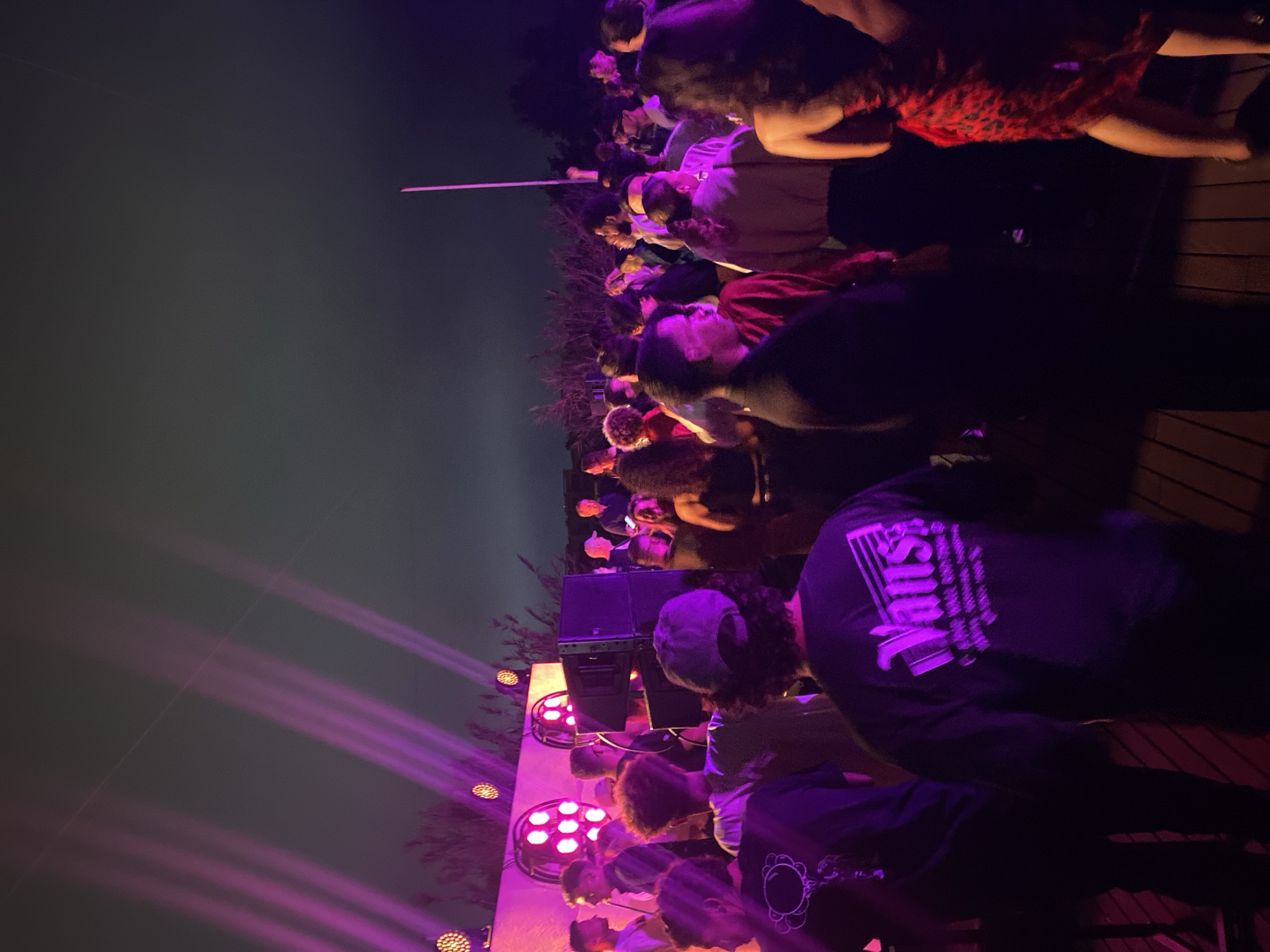 An outdoor event is underway at night in Taghazout, Morocco. Purple stage lights illuminate a crowd of people gathered on a wooden deck. A DJ booth and speakers are set up, and a white pole stands tall in the background. The atmosphere suggests a lively party or concert.