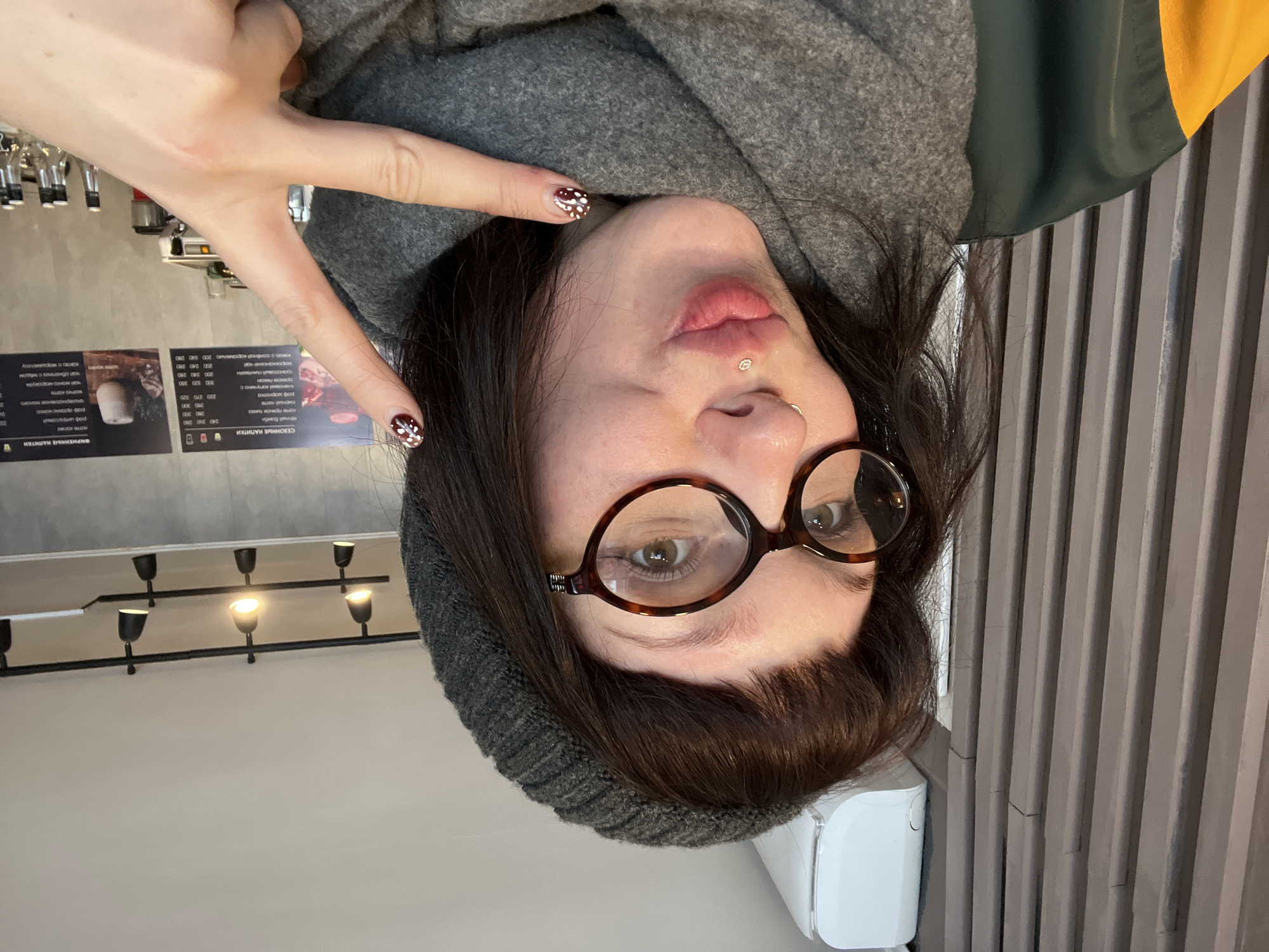 A female individual is depicted in a close-up selfie taken within a cafe in Chaikovsky, Russia. She has dark brown hair with bangs, wears a dark gray knitted beanie, tortoise-shell eyeglasses, and a gray knitted scarf. She exhibits a silver nose ring and a silver rose-shaped labret piercing. Her lips are pursed, and her light-colored eyes are directed towards the camera. She wears a dark green jacket featuring a yellow section on the left shoulder. Her right hand, adorned with dark red nails patterned with white polka dots, forms a V-sign with the index and middle fingers.
The background of the scene features a modern cafe interior. To the left, a wall with light brown vertical grooved panels is visible, topped by a partially visible white air conditioning unit. The ceiling is light-colored and fitted with black track lighting fixtures, some of which are illuminated. To the right, two dark menu boards display beverages and prices in Cyrillic text, including categories such as "СЕЗОННЫЕ НАПИТКИ" (Seasonal Drinks) and "ФИРМЕННЫЕ НАПИТКИ" (Signature Drinks). Below these boards, a counter surface is visible, holding several clear glass bottles and part of a silver espresso machine.