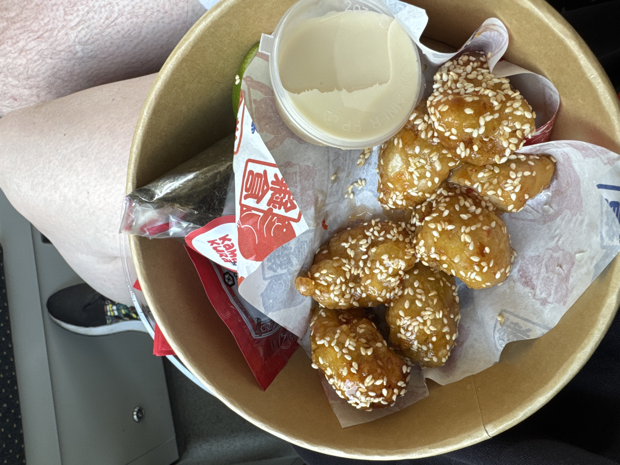 An overhead shot captures a person holding a round, light brown paper bowl filled with a snack. The primary food items are several irregularly shaped, golden-brown pieces, heavily coated in white sesame seeds and a glossy glaze, resembling fried chicken or a similar snack. These are nestled on white paper liners within the bowl.
Accompanying the main dish is a small clear plastic cup containing a light beige or off-white sauce or dip. A green lime wedge is also visible. Further within the bowl, there's a red packet with the brand name "KAMAKURA" printed in white, likely containing a condiment like chili sauce or ketchup. To the left, a clear plastic bag holds what appears to be dried seaweed or a similar crispy snack.
The background shows the person's legs and a portion of their upper thigh, indicating they are seated. A black shoe with colorful accents is visible on the lower left, resting on a grey surface. The immediate surroundings suggest an informal setting, possibly while traveling or in a casual eatery, given the disposable packaging and the visible interior paneling of what might be a train, bus, or car.
Visible text includes the brand name "KAMAKURA" on the red condiment packet. On the white paper liners, red characters, possibly Japanese or Chinese, are printed. The lid of the dip container shows "207" and partial text that might read "DINNER PP". The image captures the moment of enjoying a meal or snack. The location context is Riga, Latvia, though no specific visual cues for this city are present in the image itself.