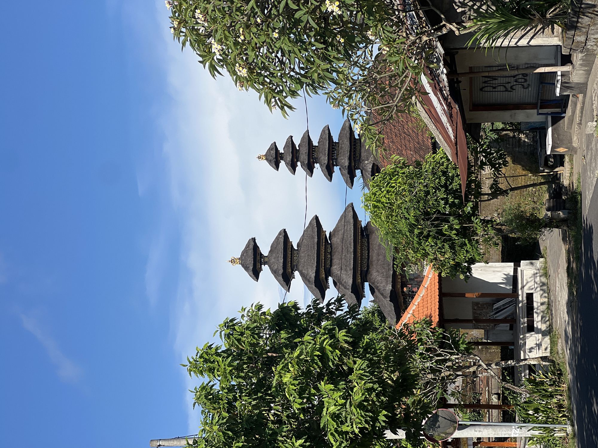 The image depicts a traditional Balinese temple structure located in Sukawati, Indonesia, set against a clear blue sky with scattered clouds. Two tiered structures, adorned with multiple progressively smaller thatched roofs, stand prominently. Lush green trees frame the temple. Adjacent to the temple, are buildings with terracotta-tiled roofs. There is a building with a rusty-looking shuttered facade and a building with an open-sided structure.