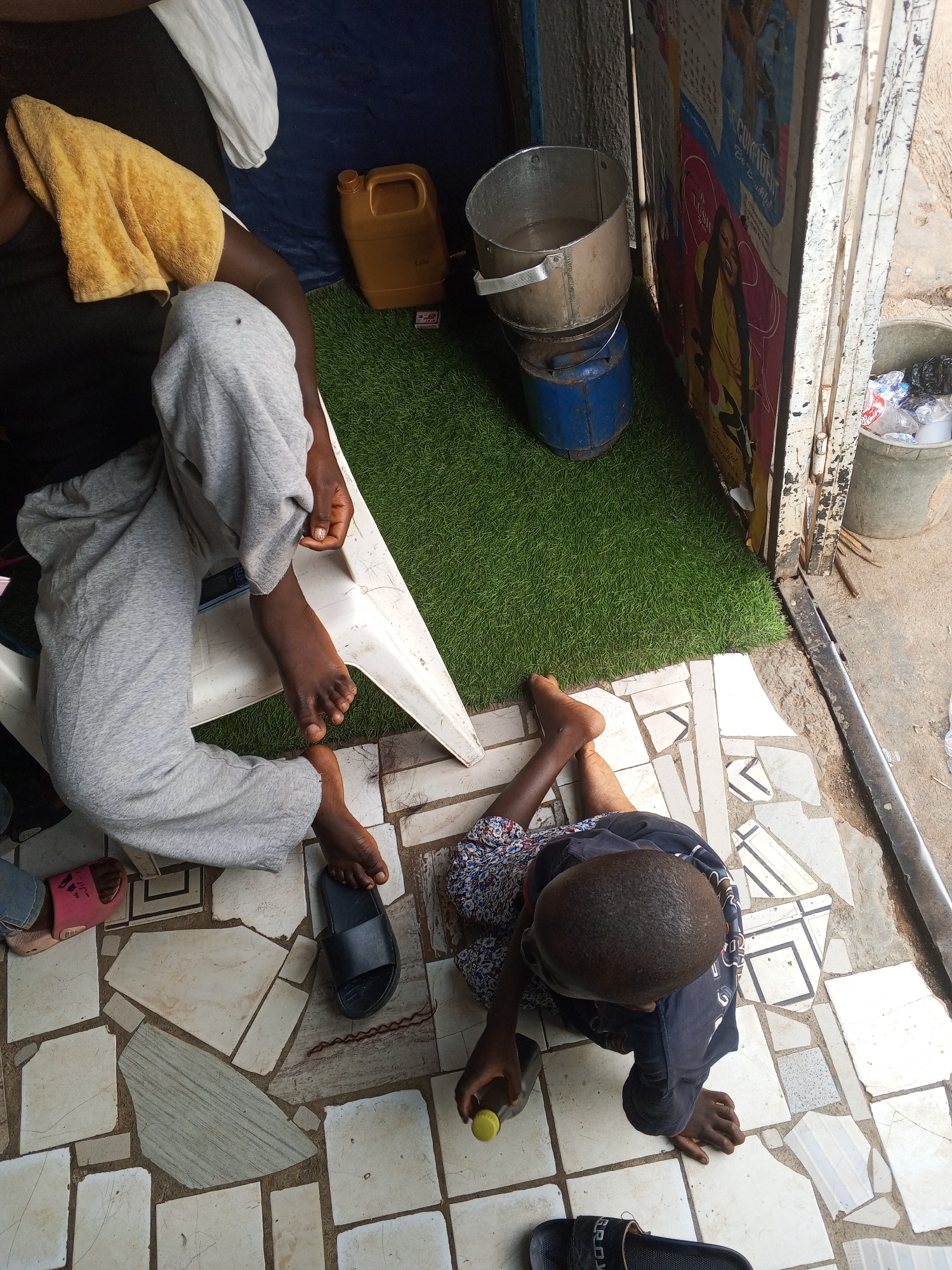A top-down shot shows a person sitting in a white plastic chair, wearing grey sweatpants and a black shirt. Another person is seated on the tiled floor. The person on the floor is wearing a dark blue top, and appears to be holding a bottle. The floor is tiled with beige tiles of various shapes. There is a patch of artificial green grass under a cooking pot, blue gas tank and yellow jerry can. There is a poster attached to the building. The location is Gwong, Nigeria.