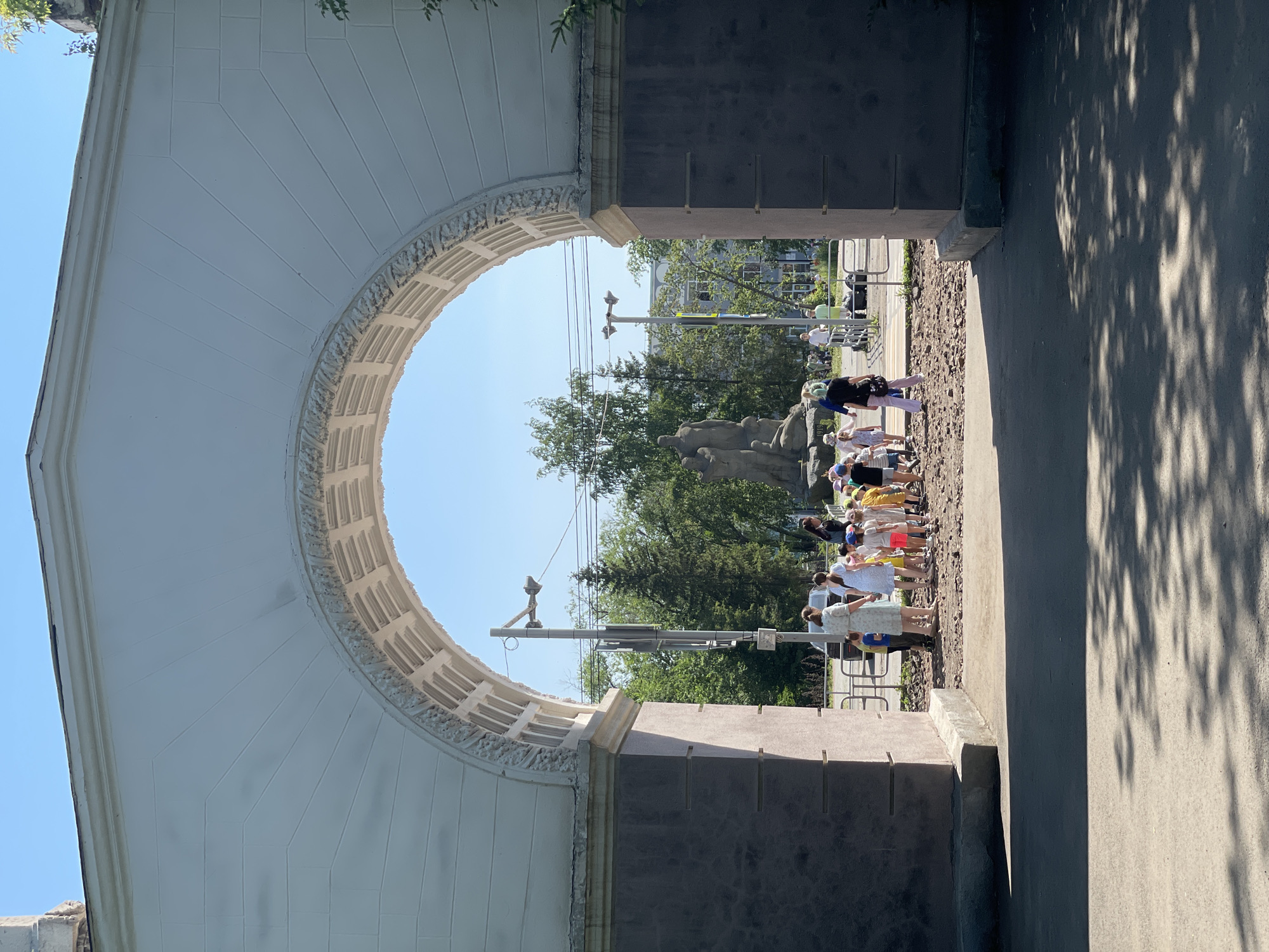 This is a medium shot, taken at eye level, of a large, white archway entrance to a park on a sunny day. Through the archway, a group of people, mostly children, are gathered near a statue. The children are dressed in casual summer clothing, and some are wearing hats. They appear to be on an outing, possibly a school field trip. The statue depicts two figures, one standing and one seated, and is located in the center of the park. Beyond the statue, trees and buildings are visible, indicating an urban setting. The archway itself is decorated with a relief pattern and its supporting pillars are dark gray. The foreground of the image shows a paved area with dappled sunlight and shadows from trees.