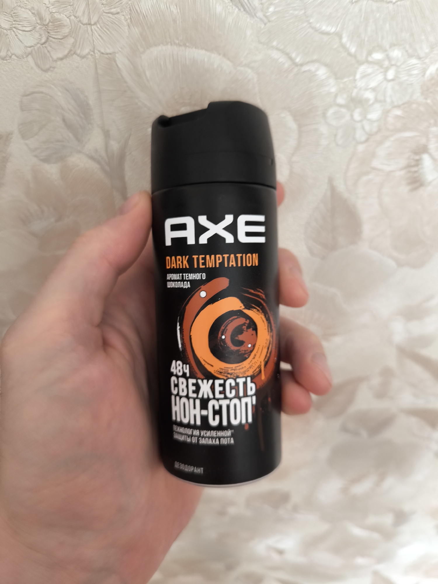 A person's hand holds a black aerosol can of Axe Dark Temptation deodorant. The can features white text that reads "AXE DARK TEMPTATION" and below that, in Russian, "AROMAT TEMNOGO SHOKOLADA" which translates to "AROMA OF DARK CHOCOLATE." There is a circular graphic with orange and brown swirls, resembling a vortex, on the can. Below this graphic, more Russian text reads "48ч СВЕЖЕСТЬ НОН-СТОП" meaning "48h FRESHNESS NON-STOP," and further text indicates "ТЕХНОЛОГИЯ УСИЛЕННОЙ ЗАЩИТЫ ОТ ЗАПАХА ПОТА" which means "TECHNOLOGY FOR ENHANCED PROTECTION AGAINST SWEAT ODOR." At the very bottom, the word "ДЕЗОДОРАНТ" (DEODORANT) is visible. The background is a light-colored, textured wallpaper with a floral pattern. The location in Gal’chino, Russia, is not discernible from the provided image.