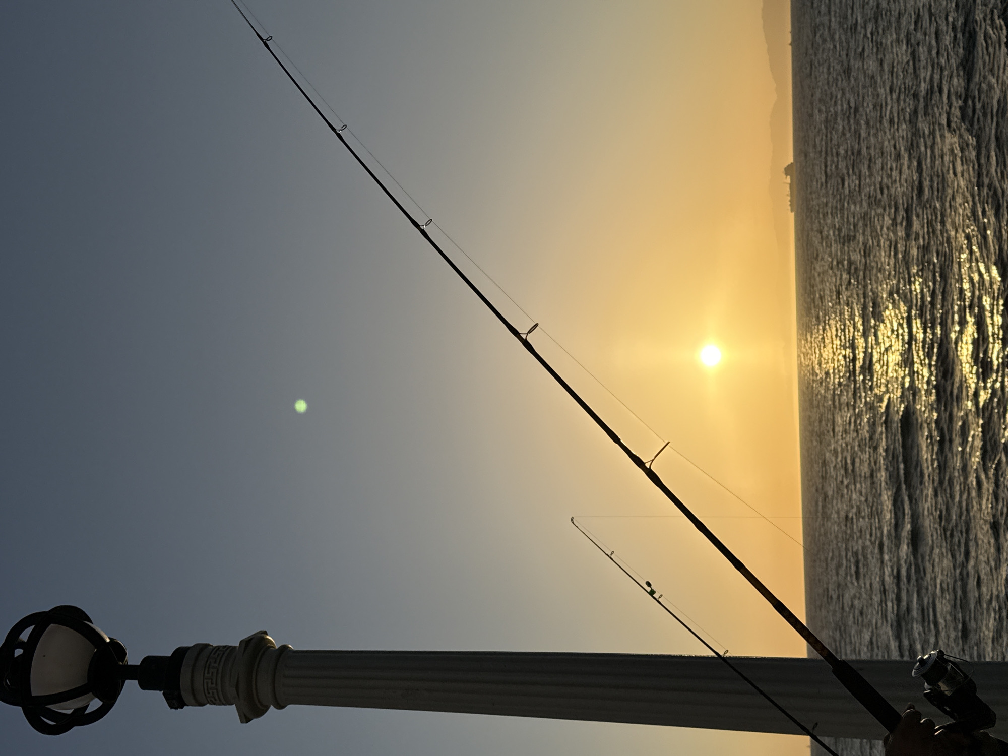 A low-angle, outdoor, full shot shows a sunset over the ocean in Manhattan Beach, California. A white lamp post with a decorative black lantern sits to the left of the frame. Two fishing rods extend diagonally across the scene from the bottom left towards the upper right. The sun, a bright orange orb, is positioned near the horizon, casting a shimmering golden reflection on the dark, rippling water. In the distance, a large cargo ship is visible on the water, silhouetted against the warm sky. The sky transitions from a clear, pale blue at the top to a gradient of orange and yellow near the horizon.