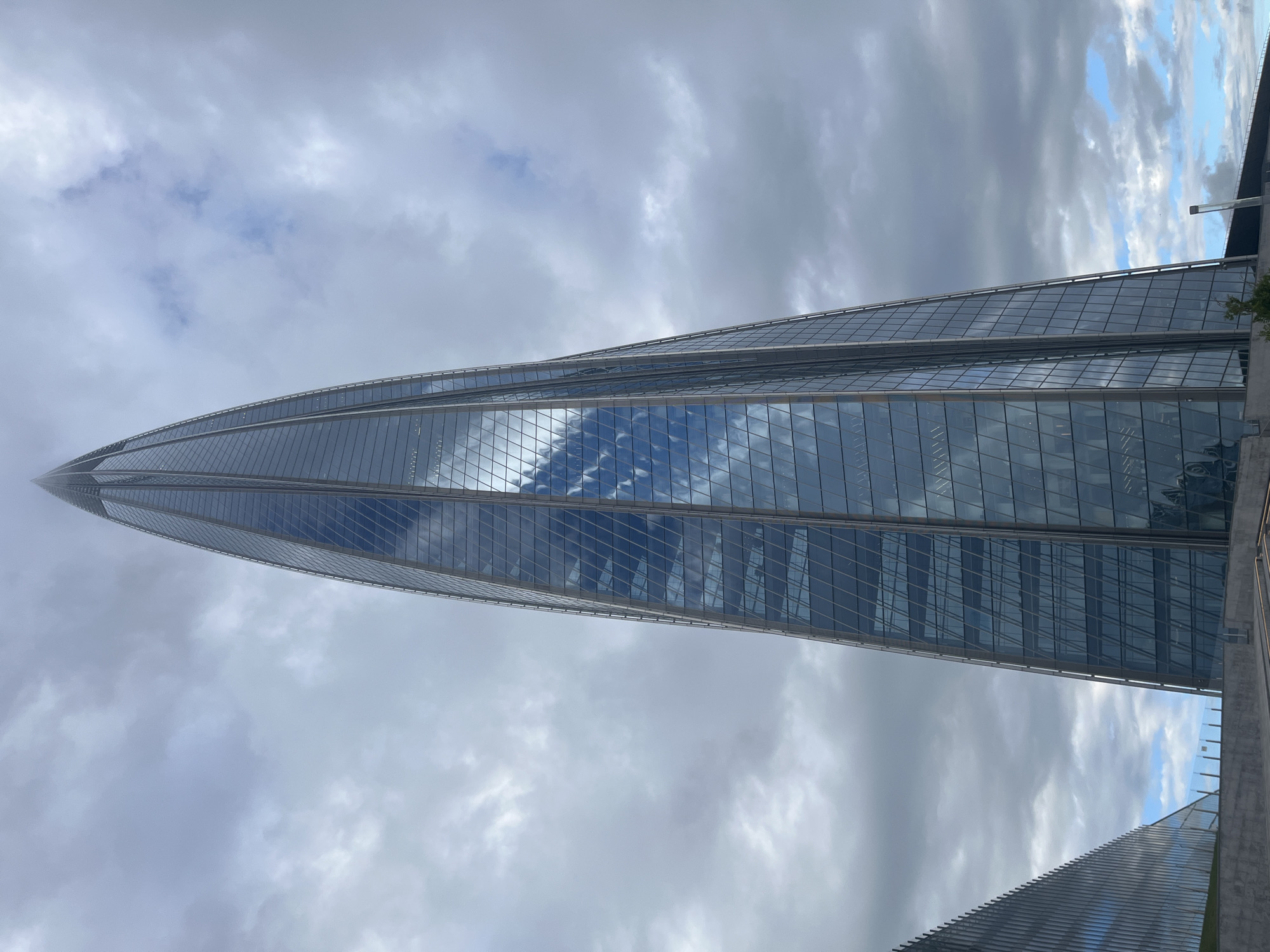 This media file shows a close-up, low-angle shot of the Lakhta Center skyscraper in Saint Petersburg, Russia. The skyscraper's distinctive twisted, glass facade dominates the frame, reflecting the cloudy sky above. The building tapers to a point and appears to pierce through a layer of fog or low-lying clouds near its base.
The sky is overcast with a mix of dark gray and lighter, patchy clouds, suggesting either early morning, late afternoon, or a generally cloudy day. Patches of blue sky are visible, indicating some breaks in the clouds. The lighting is diffused, consistent with cloudy weather.
The scene captures the immense scale and modern architectural design of the Lakhta Center. There are no people or specific activities visible, focusing solely on the imposing structure and its interaction with the atmosphere. The building's reflective glass panels mirror the sky, creating a sense of depth and emphasizing the building's height. The image provides a dramatic perspective of the skyscraper against a moody sky, highlighting its iconic silhouette. There is no visible text in the image.