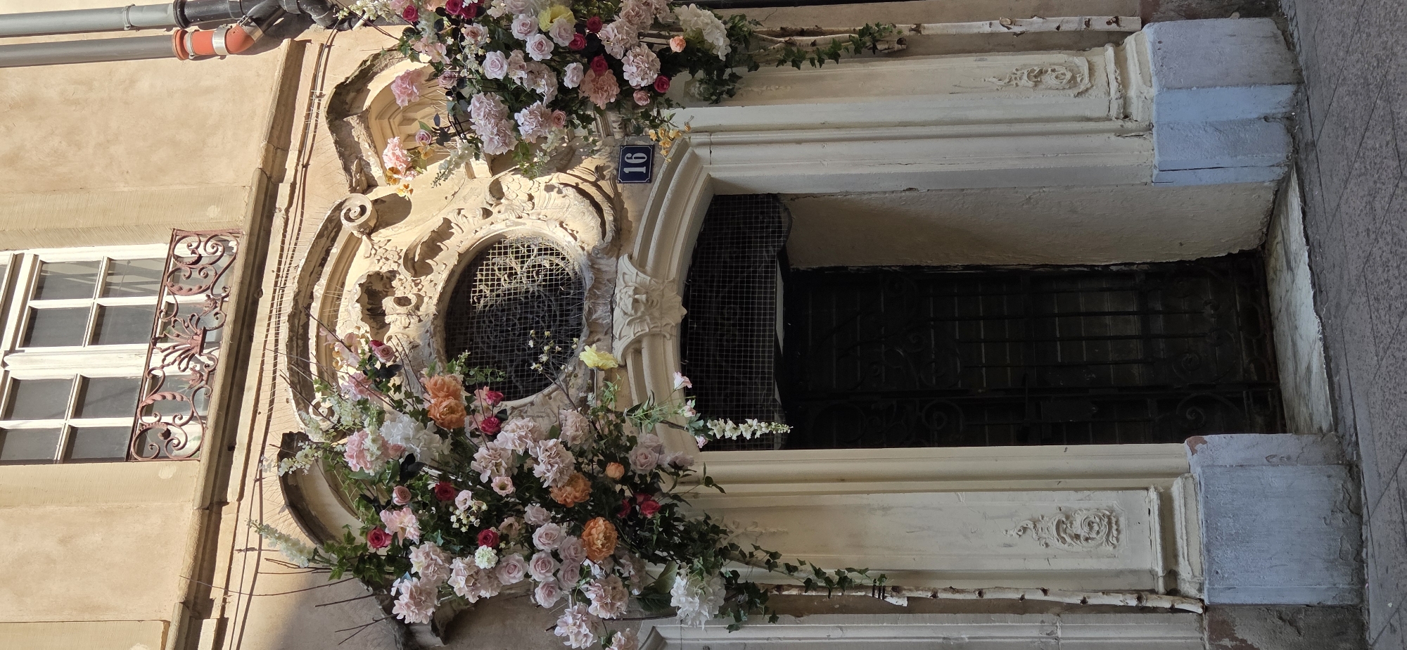 The image shows the ornate entrance to a building in Strasbourg, France, decorated with large floral arrangements. The building facade is made of a light-colored stone or stucco. Above the entrance, there is a decorative window with a metal grille and a carved stone surround featuring ornate details. A wrought-iron railing is visible in front of a window higher up on the facade. On either side of the entrance arch, there are substantial floral displays composed of pink, white, and peach-colored flowers, with greenery. A dark metal door with intricate scrollwork is visible behind a wire mesh covering the upper part of the doorway. A blue rectangular sign with the white number "16" is mounted on the wall to the right of the door. The ground in front of the entrance is paved with cobblestones. The overall scene suggests a building with historical architecture, possibly decorated for an event or seasonal display.