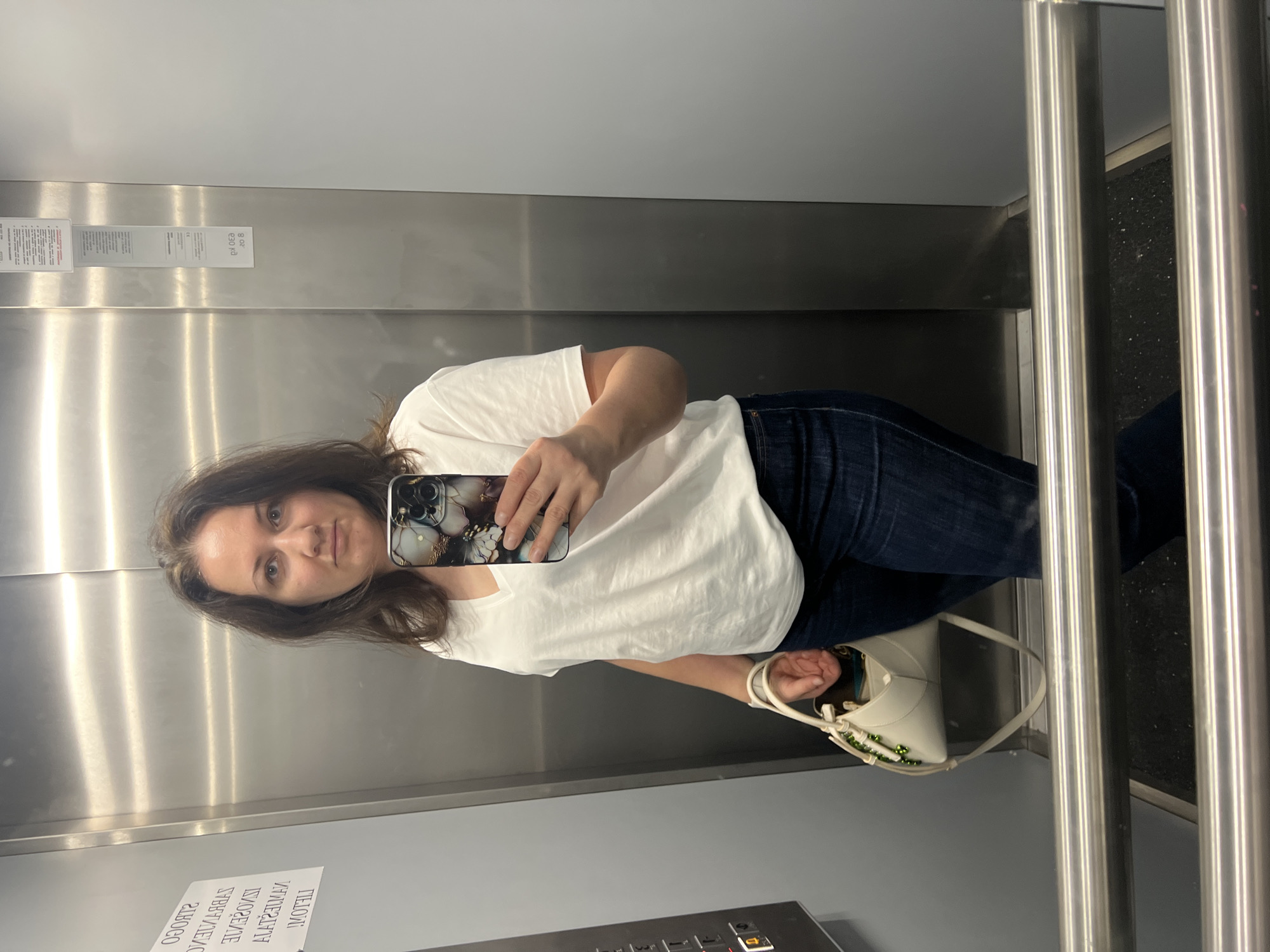 A medium-shot photograph shows a woman facing the camera in what appears to be an elevator, judging from the metal walls and the presence of vertical metal bars. She is holding a mobile phone up to take a selfie. The woman is wearing a white t-shirt and dark jeans. She has shoulder-length brown hair. A light-colored handbag is hanging from her arm. The elevator's walls appear to be made of brushed metal. There's an array of elevator buttons at the bottom. A mirrored surface reflects the scene. The location of the image is not discernible from the available information. Budva, Montenegro, is not identifiable from the image.