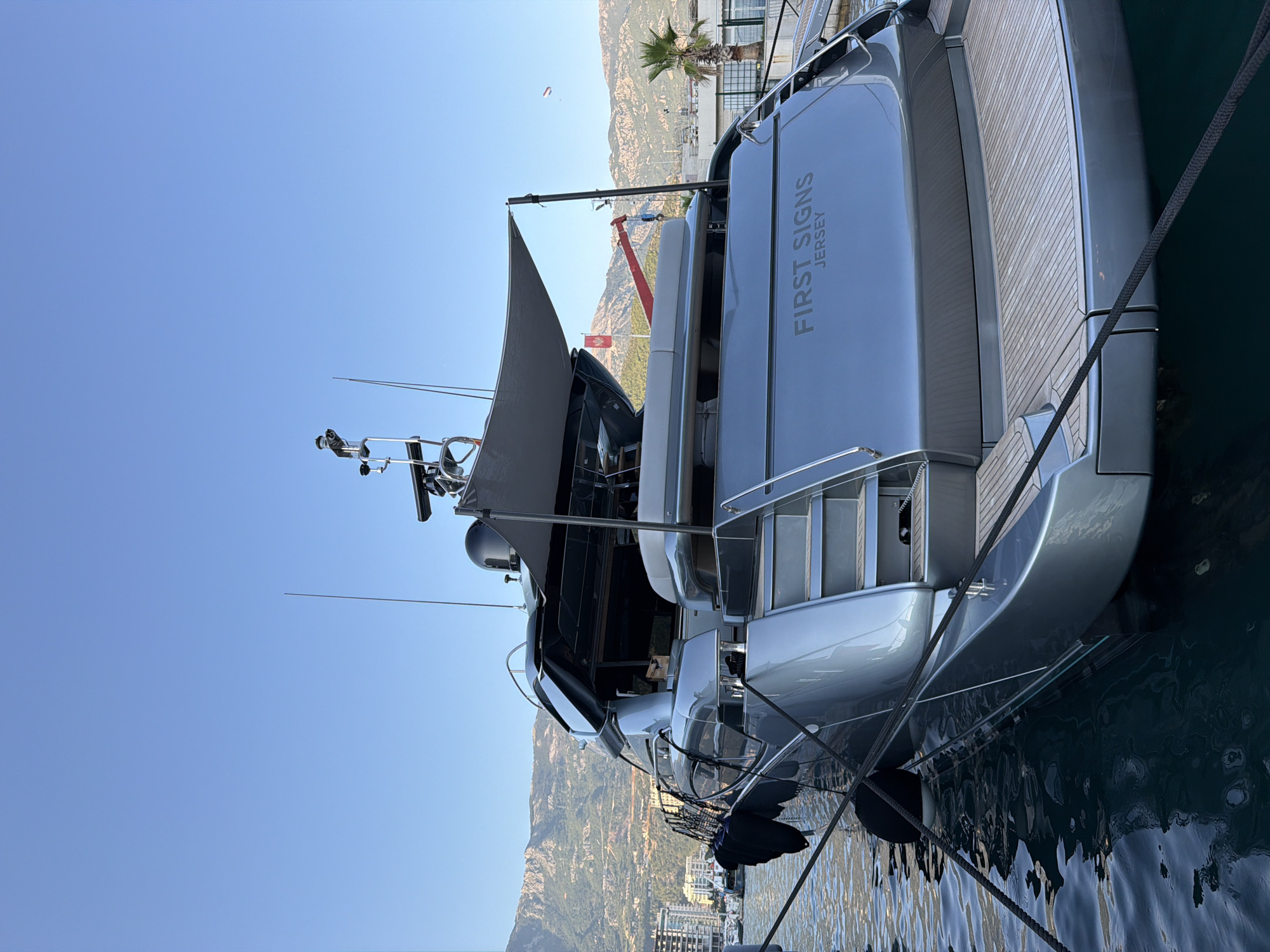 The image shows the stern of a grey luxury yacht, named "FIRST SIGNS JERSEY", docked in the marina of Budva, Montenegro. The yacht is moored with black ropes, and black fenders are attached to its side. The stern features a teak deck and a set of stairs leading up to a higher level. In the background, the cityscape of Budva is visible, with buildings nestled against a backdrop of green, mountainous terrain under a clear blue sky. A palm tree is visible on the right side of the image, and a red flag is flying from a pole on the hillside. The water in the marina appears calm and dark.