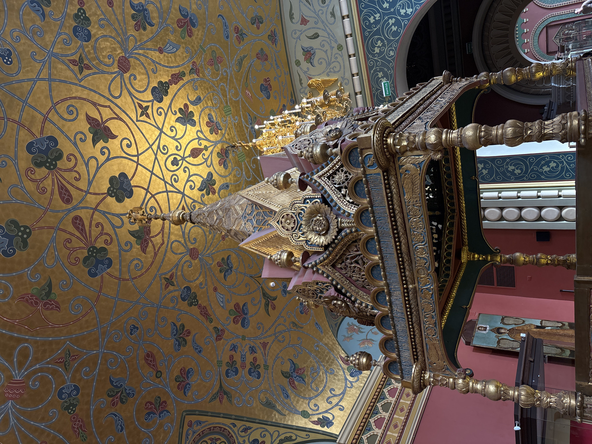 The image shows an interior view of a building, likely located in Moscow, Russia, as indicated by the Russian text "выход" (exit) on a green sign. The perspective is tilted upwards, showcasing the ornate ceiling and a decorated structure within the building.
Ceiling: The ceiling is predominantly gold, adorned with a complex floral pattern in blue, green, and red hues.
Structure: A golden ornate structure dominates the view, featuring intricate carvings, patterns, and potentially, lighting fixtures.
Walls: The walls are painted in shades of red and blue, with patterned borders along the edges. An icon is visible hanging on the wall.
