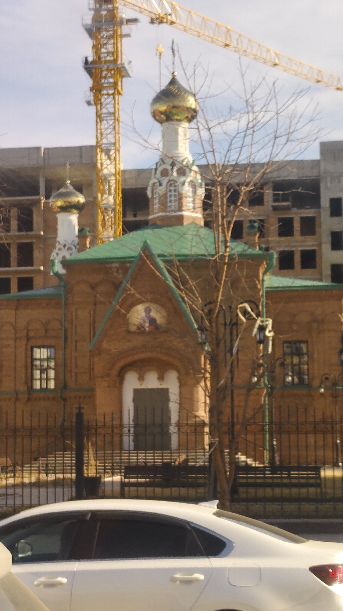 A daytime shot of an orthodox church in an urban environment. The church is made of red brick, with a green roof and golden domes. In the background, a yellow crane and a building under construction are visible. In the foreground, a white car is partially visible. A black wrought-iron fence separates the church from the street. A bare tree stands to the right of the church. The location is Unknown, Unknown.