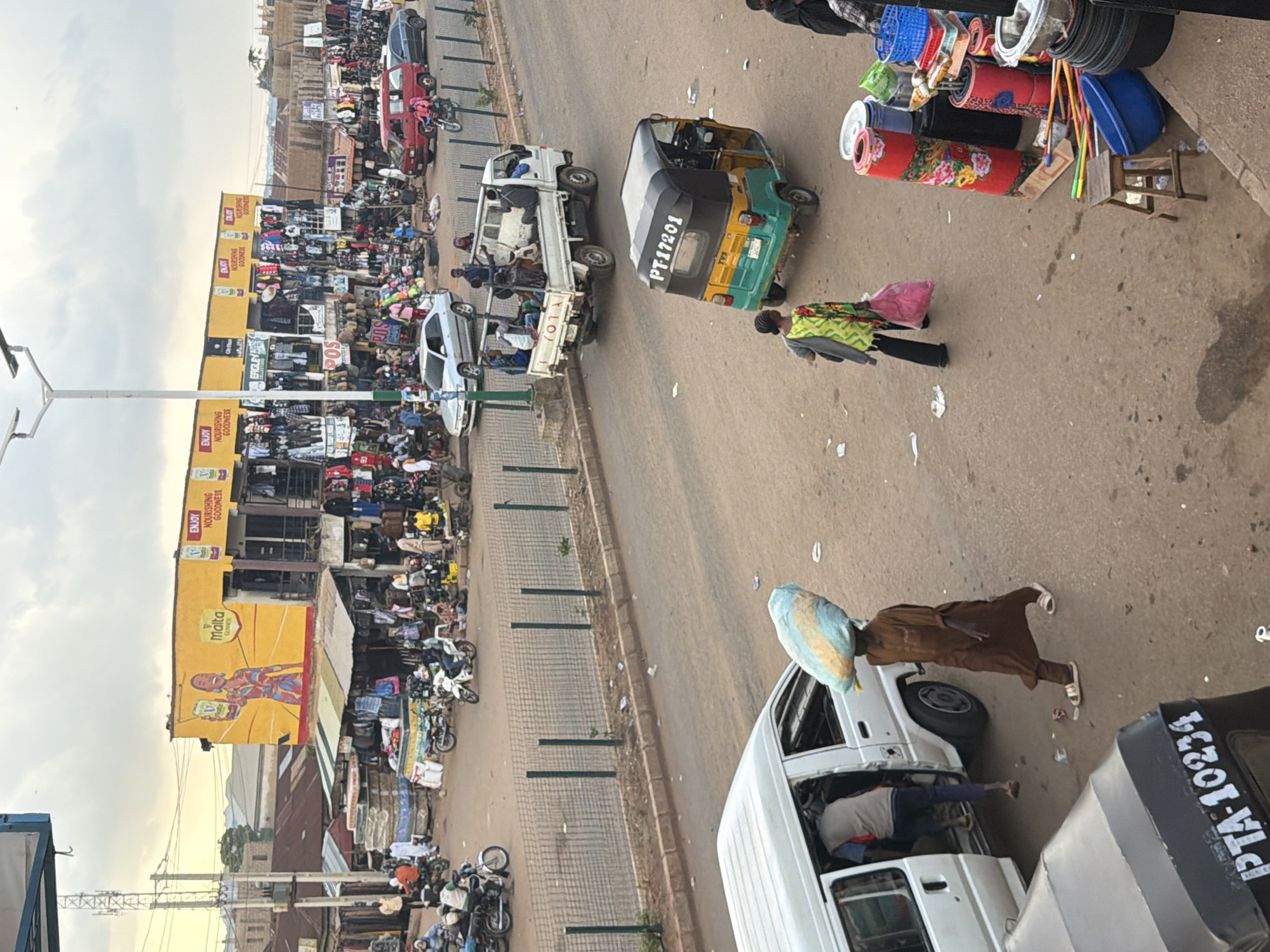 This is an outdoor, eye-level medium shot of a bustling street market scene in Bukuru, Nigeria. The image is dominated by a colorful yellow building with advertisements for "Malta Guinness" and "Enjoy Nourishing Goodness". Below the building, market stalls are set up with clothing and various goods displayed.
In the foreground, a white van is parked with its door open, and a person in a brown robe is walking away from it, carrying a large, light blue and yellow object on their head. Another person, dressed in a dark garment, is visible inside the van. To the right, a green and yellow auto-rickshaw with the license plate "PT-17201" is visible. In the middle ground, several vehicles, including cars and motorcycles, are present, along with people walking and interacting in the market. A wire fence separates the market area from the road. The background shows more buildings and vegetation under a cloudy sky. The overall impression is one of a lively commercial hub.
