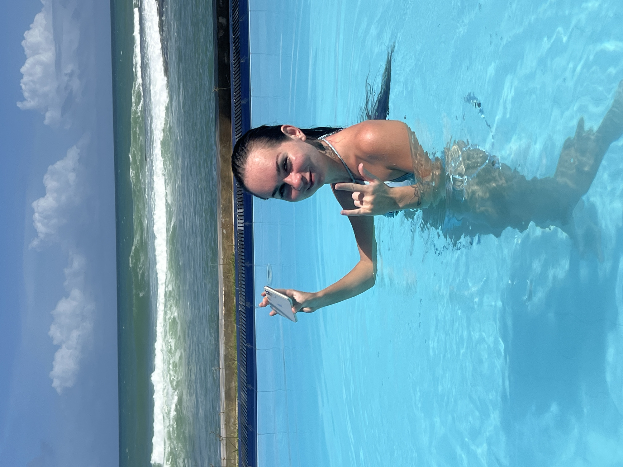 In the city of Goviyapana, Sri Lanka, a young woman is posing in a bright blue swimming pool. She is partially submerged, with her head and shoulders visible above the water. Her wet, dark hair is down her back. She is holding a smartphone in her right hand, displaying it towards the camera. Her left hand is raised, with her index and pinky fingers extended in a "rock on" gesture. She is wearing a light-colored swimsuit and a necklace. The pool's blue tiles are visible along the edge. Beyond the pool, the turquoise ocean with white-capped waves under a partly cloudy blue sky can be seen, suggesting a coastal location. The overall atmosphere is one of leisure and enjoyment.