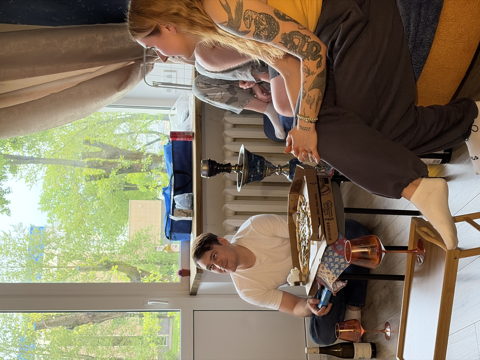 The interior scene depicts a social gathering centered around a pizza meal. Three individuals are visible in the composition. A fair-skinned, light-haired woman with numerous arm tattoos sits on a chair and observes the scene. A person is seen in the background. In the foreground, a fair-skinned man in a white t-shirt holds a small, blue electronic device. A pizza box is on the table, with several slices missing. Two wine glasses and a bottle of wine rest near the table. A hookah is on the table. Outside the window, foliage suggests a tree-lined outdoor environment.