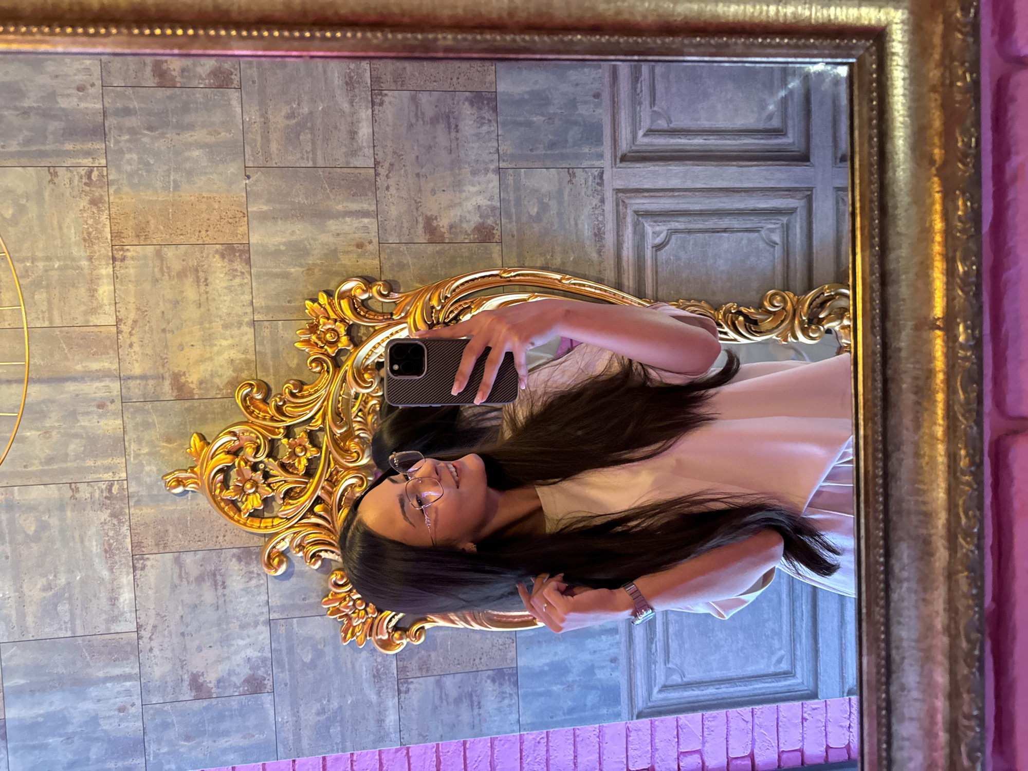 A young woman with long, dark hair and clear-framed glasses is depicted in a mirror selfie, with the image rotated 90 degrees clockwise. She wears a light-colored top and a silver-toned watch on her left wrist, and holds a smartphone with a textured dark case in her right hand to capture the reflection. Her expression is pleasant, with a slight smile.
The mirror features an ornate, golden, baroque-style inner frame, and is surrounded by a larger, simpler golden outer frame. Reflected in the mirror behind the woman is an interior wall composed of rectangular panels with a distressed, rustic finish in shades of grey and brown. To the right, a grey panelled door is visible. Below the ornate mirror frame, a section of a wall with pink/purple brick-like texture is reflected. A partial golden, curved metallic structure is also visible on the left side of the reflection.
The activity taking place is the woman taking a mirror selfie. The setting is an indoor space, likely a cafe, restaurant, or a stylish home in Sofia, Bulgaria, given the decorative elements and unique wall textures. No text is visible in the image.