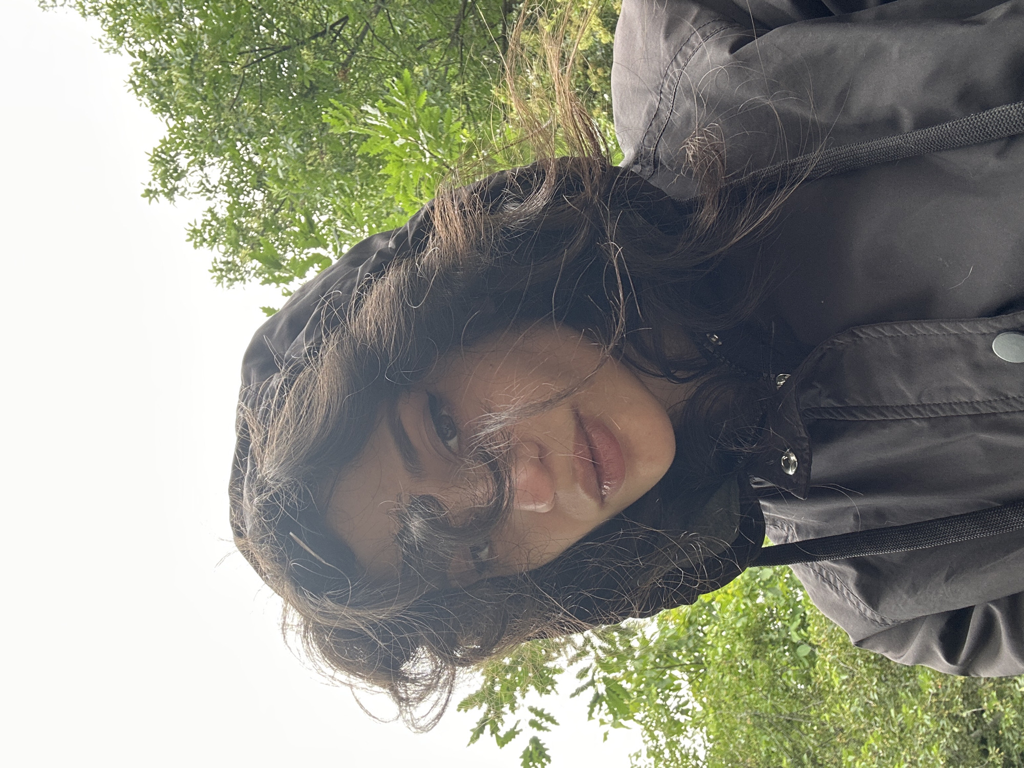 A low-angle, selfie-style shot features a young woman looking up at the camera, her face framed by dark, wavy hair that is being blown by the wind. She is wearing a dark, hooded jacket, with the hood pulled up around her head. The background consists of lush green trees and a bright, overcast sky, suggesting an outdoor setting, possibly a park or wooded area within the city of Sofia, Bulgaria. The lighting is diffused, indicating a cloudy day. The woman's expression is neutral, with her gaze directed intently at the viewer. The focus is on her face and upper torso, with the natural environment serving as a blurred backdrop.