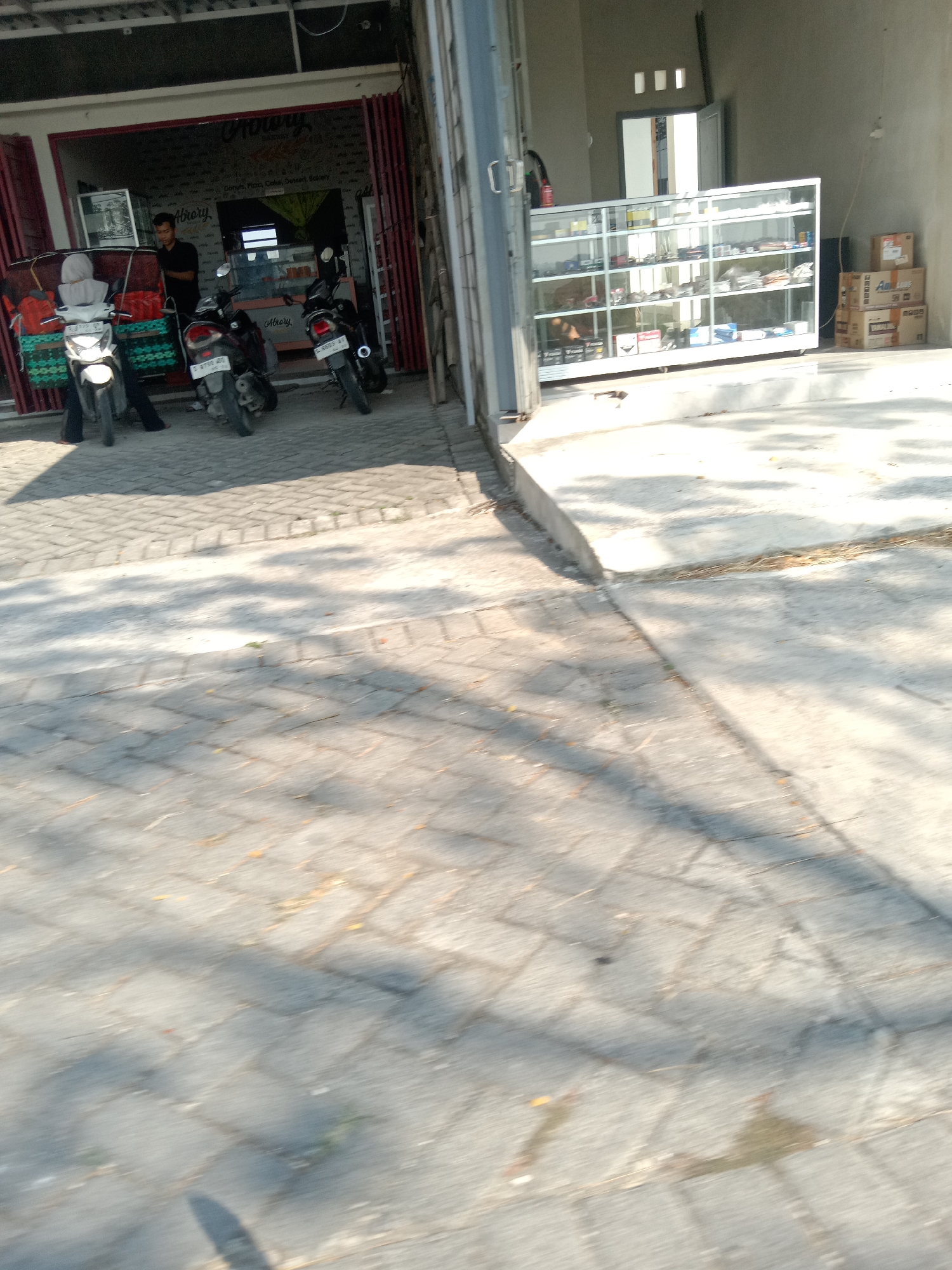 The image captures a street scene in Kemamang, Indonesia, featuring a bakery and a display case. In the foreground, a paved area with interlocking bricks shows dappled sunlight and shadows. To the left, a bakery with a red accordion-style door is partially open, revealing the interior. A man in a black shirt stands near the counter inside. Three motorcycles are parked in front of the bakery, two of them appearing to be the subject of recent activity. A person wearing a grey hooded jacket sits on one of the motorcycles. In the middle ground, a glass display case filled with various items is positioned outside a building to the right. The case has multiple shelves, and the items on display are not clearly identifiable but appear to be packaged goods. Behind the display case, boxes are stacked, some with labels. The background shows a building with a grey wall and a doorway. The overall scene suggests a commercial area with outdoor elements.