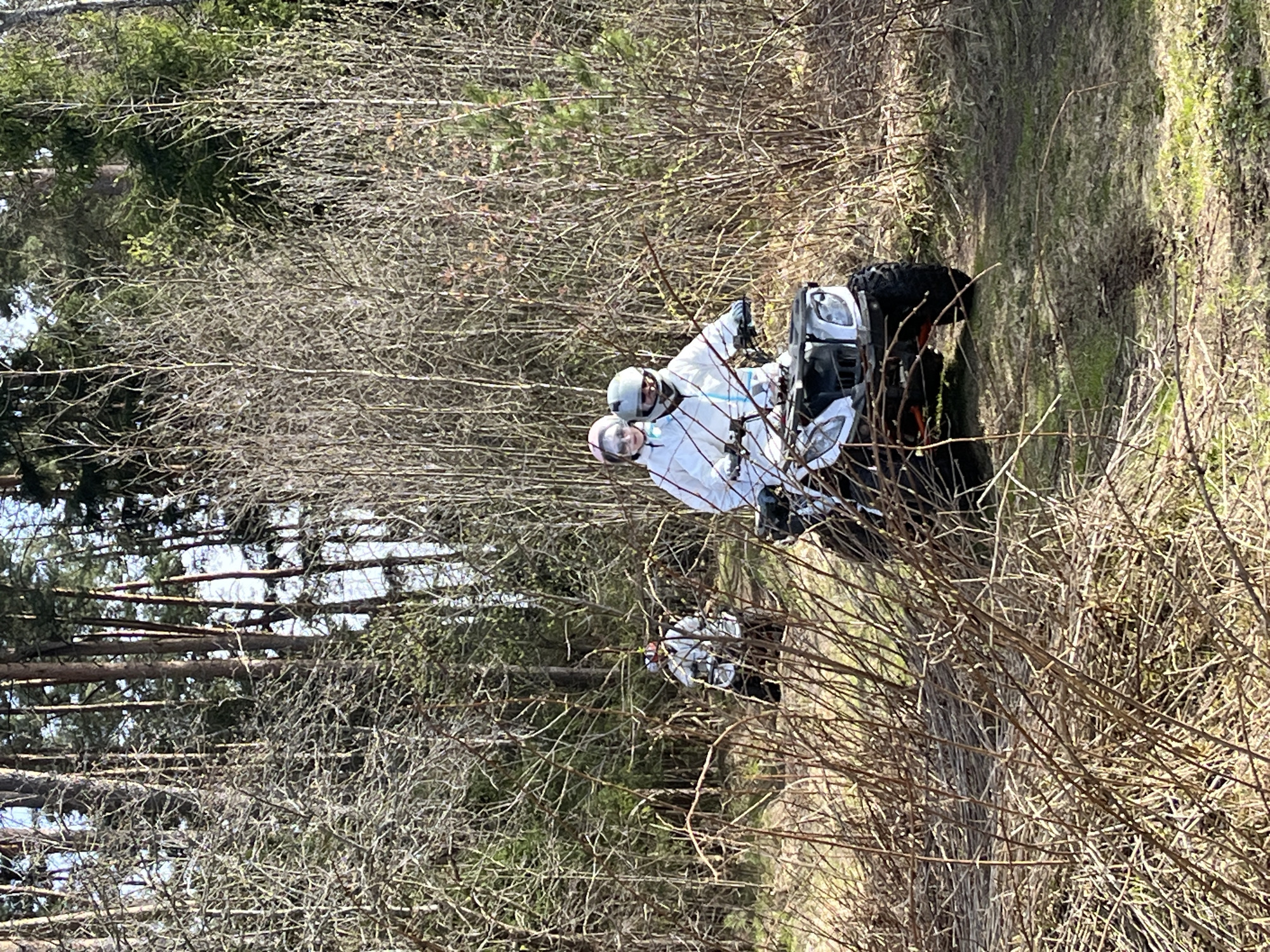The image depicts two individuals on an ATV riding on a dirt trail through a forest in Razdolye, Russia. The individuals are wearing helmets and white jackets. The surrounding environment consists of trees and bare bushes. The ATV is partially obscured by the vegetation.