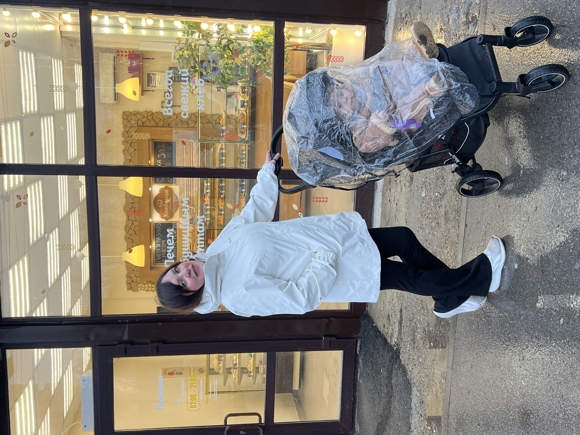 A woman wearing a white jacket and black pants stands outside a building in Perm, Russia, identified by a sign in Russian. She is holding a black stroller with a clear plastic cover, revealing a baby or child inside. The building behind her appears to be a bakery, judging from the signs mentioning "fresh bread" and "pies." The area outside looks slightly wet, suggesting it might have recently rained. The time on the door indicates the operating hours from 07:00 to 21:00.
