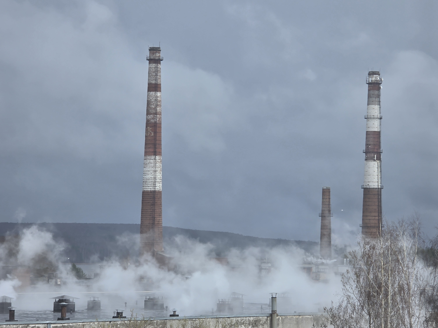 The image depicts an active industrial complex, likely a thermal power station, under a heavily overcast, grey sky. The scene is set in Chaikovsky, Russia.
Dominating the mid-ground are two tall, red and white striped brick chimneys. The chimney on the left features visible Cyrillic text: "ТЭС" (TES, likely standing for Thermal Power Station or Thermal Electric Station) and below it, "1973", possibly indicating its year of construction or a significant date. A smaller, plain brown chimney is visible between and behind the two main chimneys, with another partially visible further back.
A significant amount of white steam or smoke billows upwards from the lower industrial structures, obscuring much of the facility at ground level. In the immediate foreground, the top of a concrete wall or building roof with several ventilation structures is visible, also partly enveloped in steam. To the far right, a cluster of bare-branched trees, suggesting a late autumn or winter season, adds to the starkness of the landscape. In the distance, a line of forested hills can be seen under the dark, heavy clouds. The overall impression is one of active industry on a cold, dim, and cloudy day.